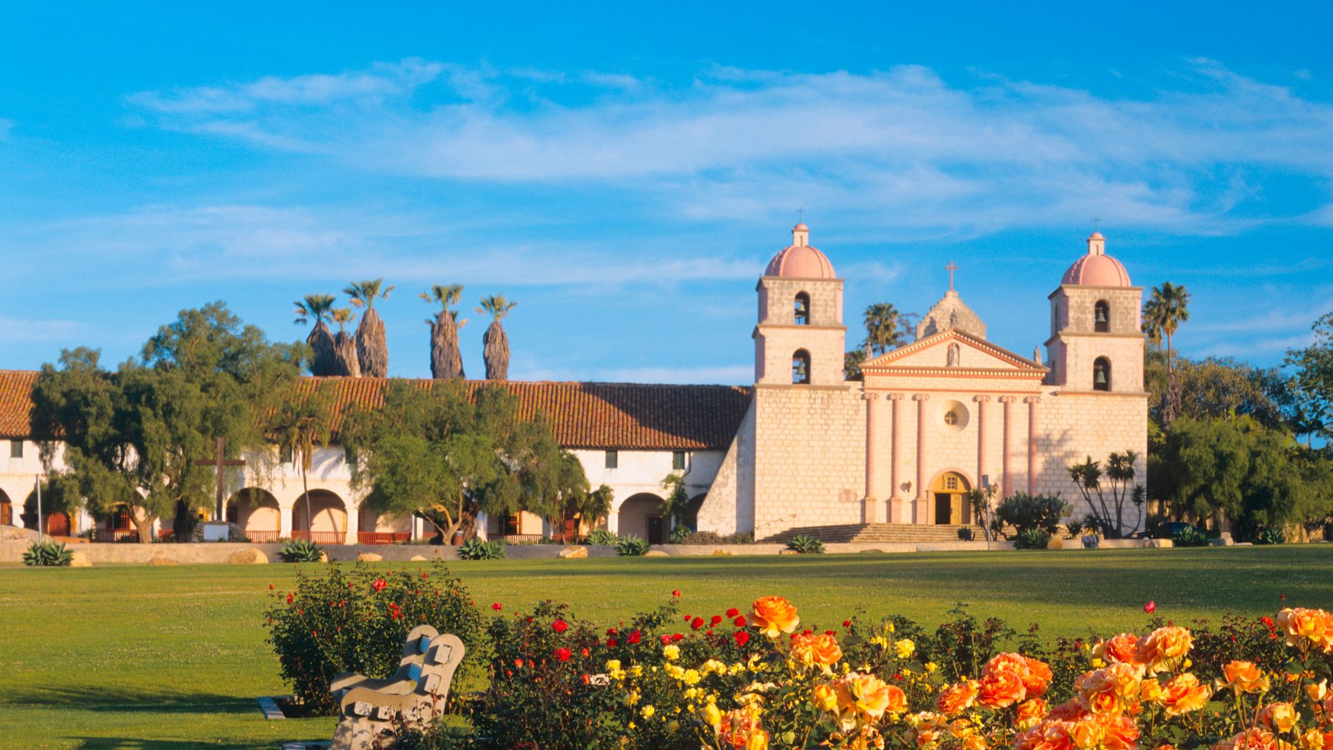 A wide shot of Mission Santa Barbara in California, featuring its distinctive neoclassical façade with twin bell towers, set against a bright blue sky. In the foreground, a vibrant green lawn is bordered by colorful flowerbeds with blooming orange and red flowers. Palm trees are visible behind the mission's structure.