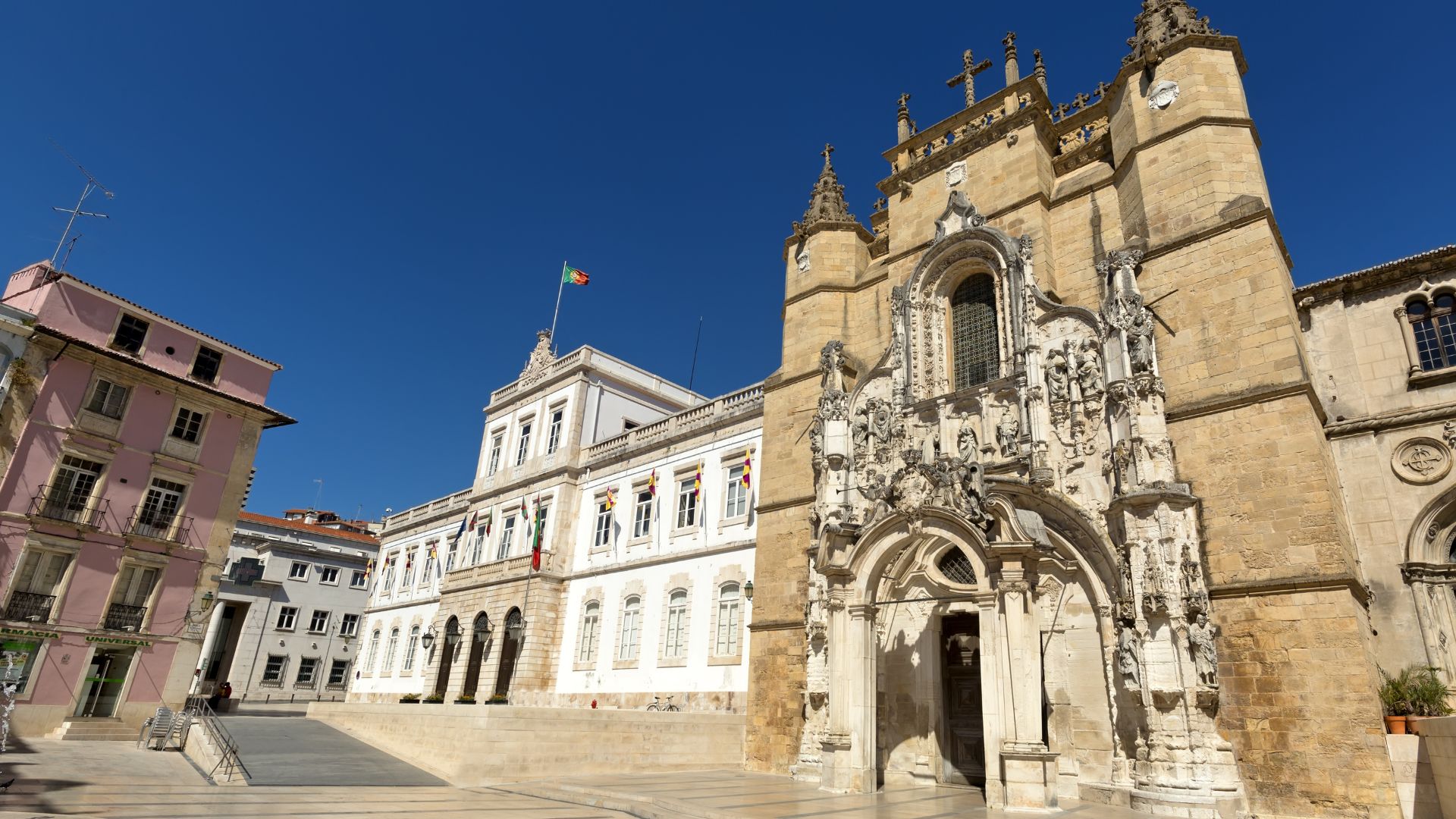 A wide-angle view of the Monastery of Santa Cruz in Coimbra, Portugal, featuring its ornate stone facade and entrance, with a clear blue sky overhead and other buildings and a paved square visible in the foreground and to the left.