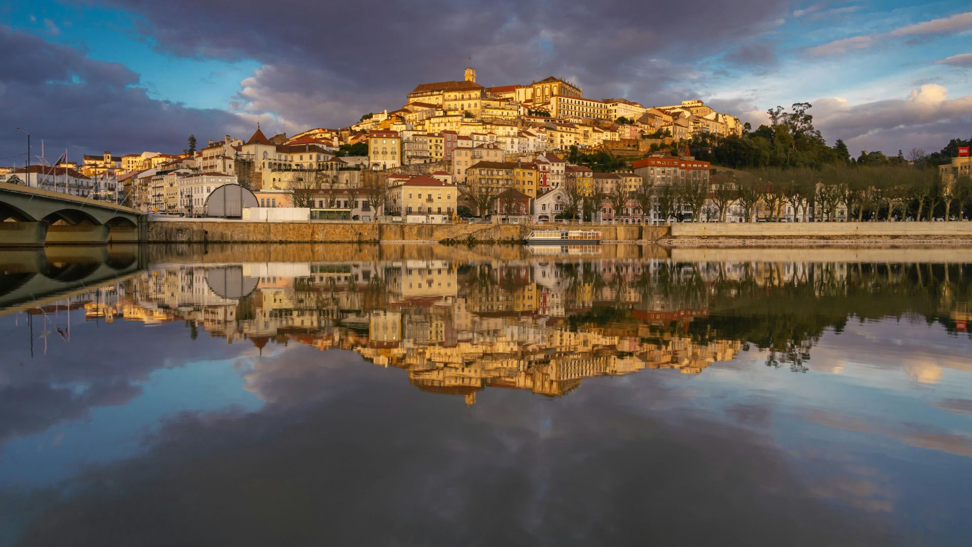 The historic city of Coimbra, Portugal, bathed in the warm glow of sunset, is reflected in the tranquil waters of the Mondego River. The city's iconic buildings, including the prominent University, ascend the hillside, creating a picturesque and timeless scene. 