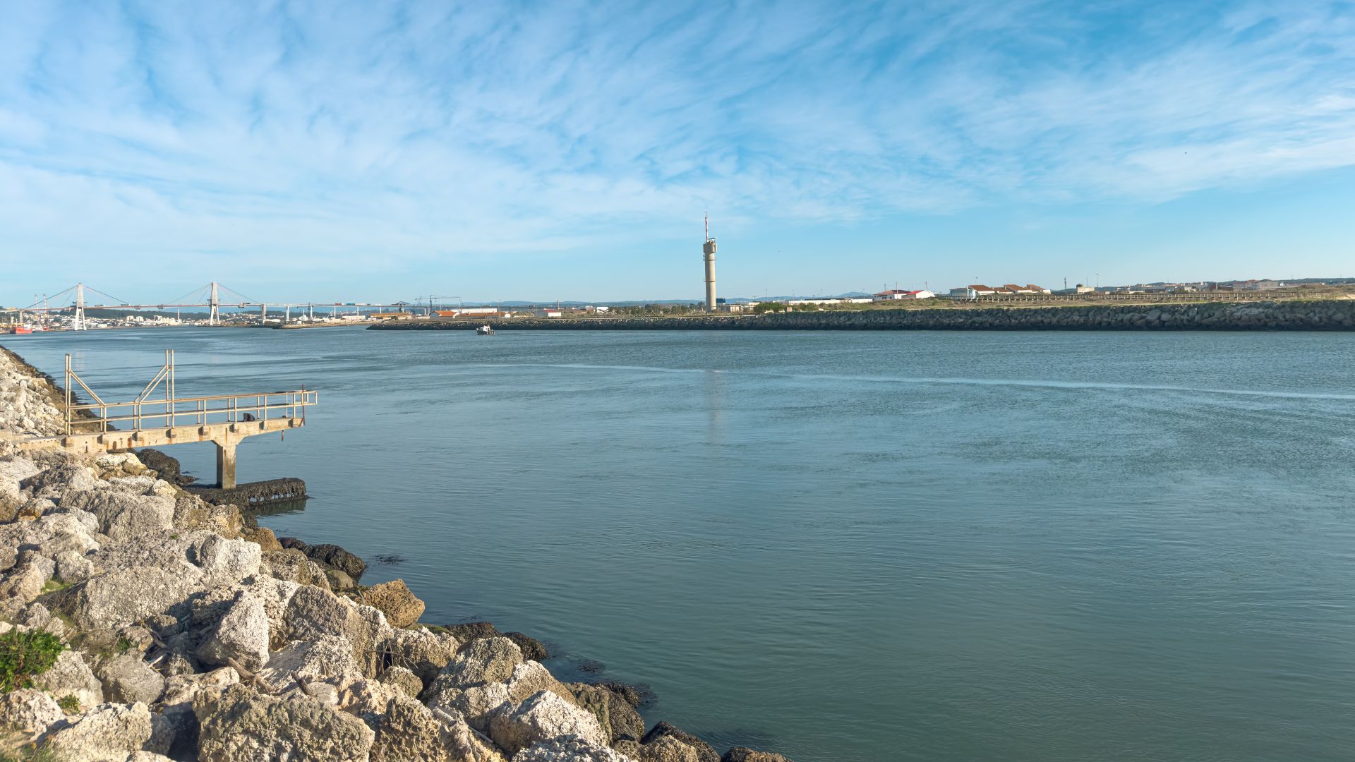 A wide river, likely the Mondego, flows under a bright blue sky with scattered clouds. On the left, a rocky shoreline with a small pier extends into the water, while the distant right bank shows urban development and a prominent tower.