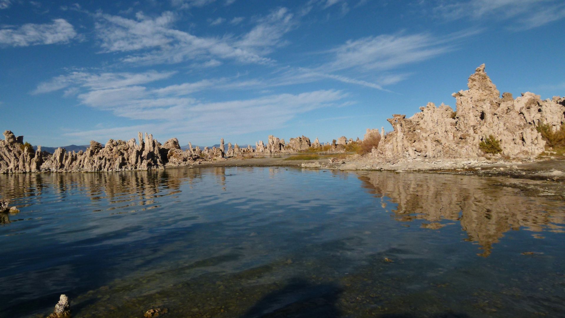 A panoramic view of Mono Lake in California under a bright blue sky with scattered clouds. The calm, reflective water in the foreground mirrors the dramatic, light-colored tufa towers rising from the water and along the shoreline in the midground and background, creating a unique and striking natural landscape.