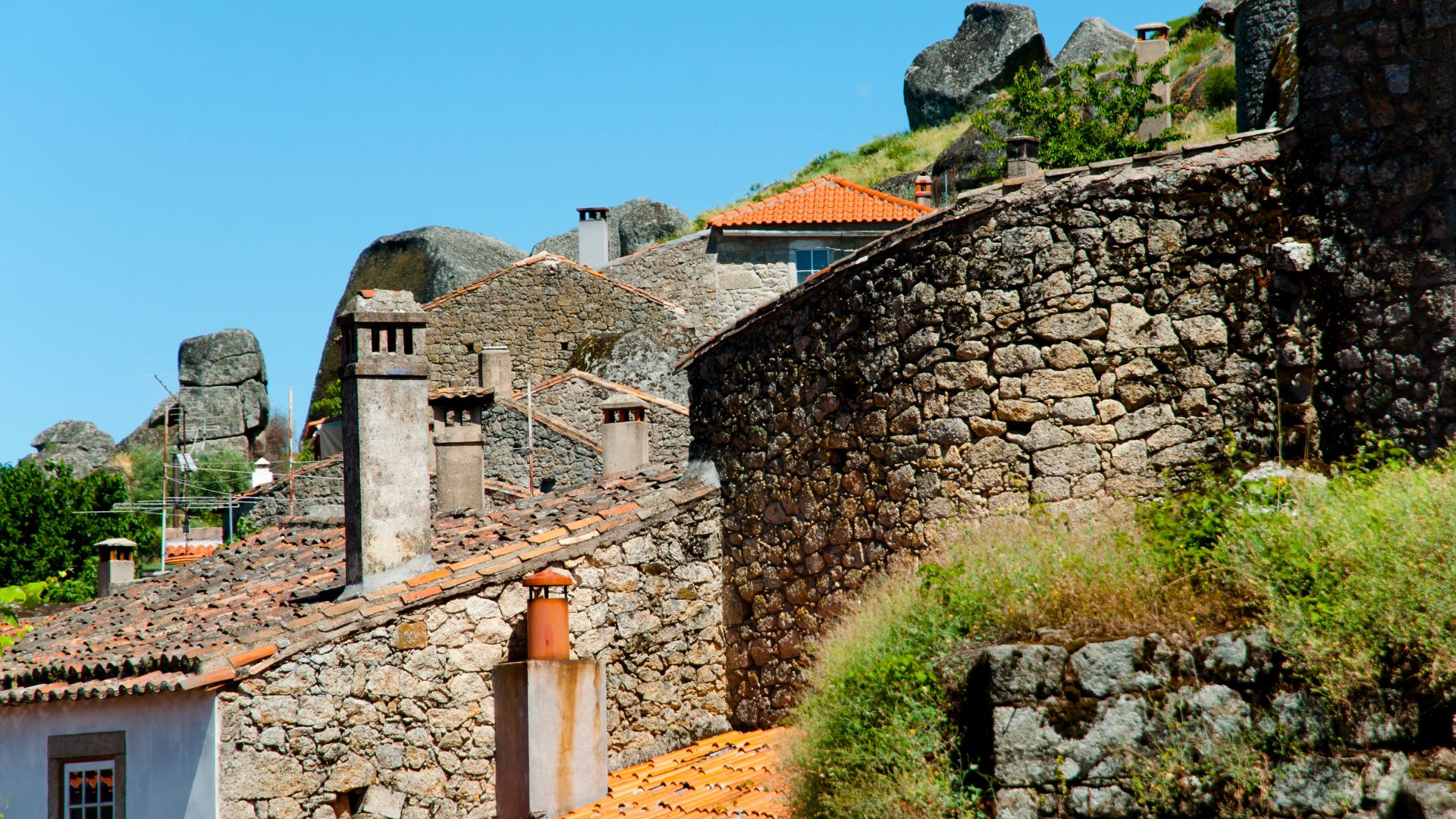 A view of the unique village of Monsanto, Portugal, showcasing its distinctive stone houses built among large granite boulders, with tiled rooftops and chimneys visible against a rocky hillside under a clear blue sky. 