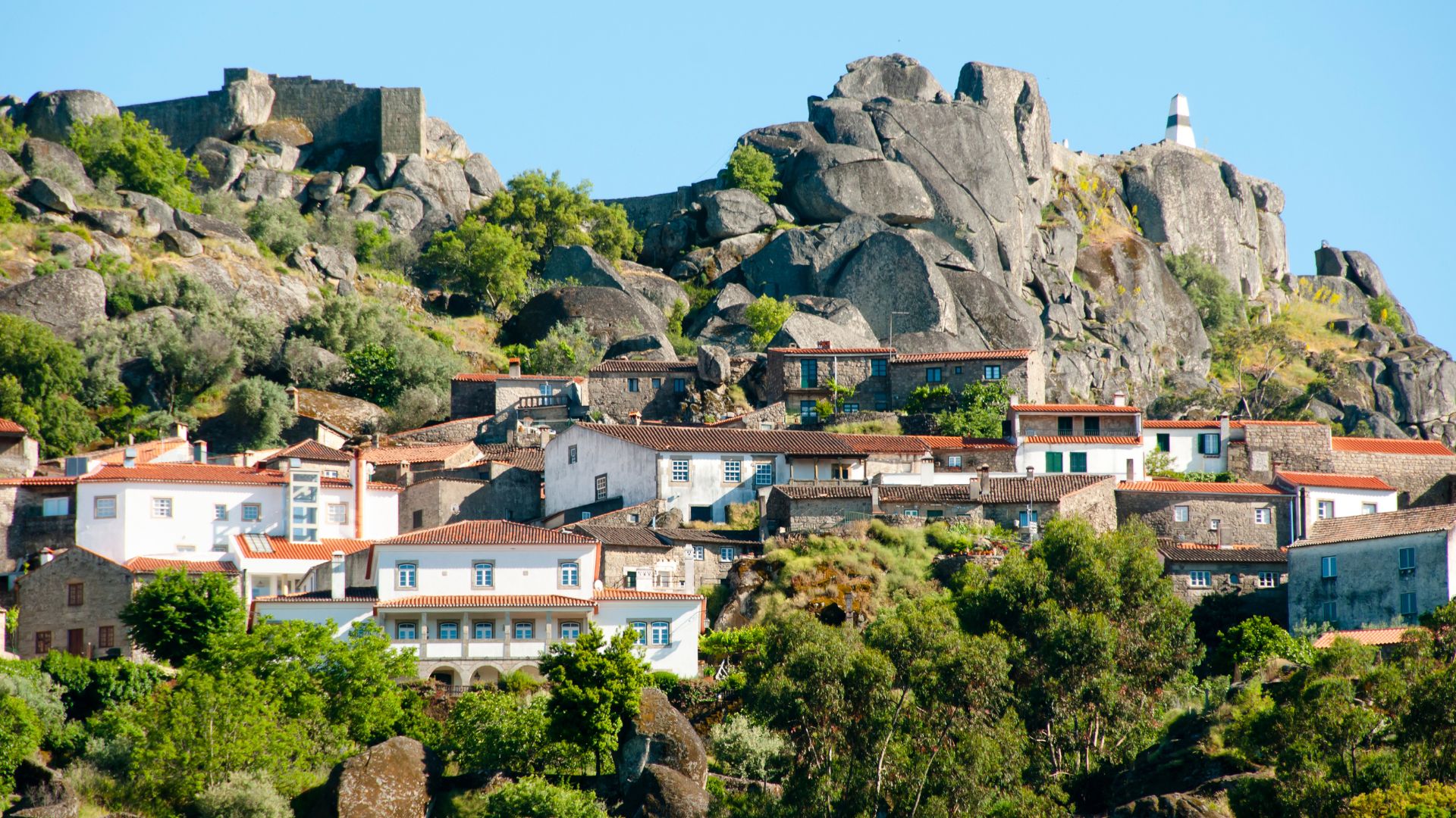 A scenic view of the historic village of Monsanto, Portugal, characterized by its unique architecture with houses nestled among massive granite boulders and a medieval castle ruin visible on the hilltop.