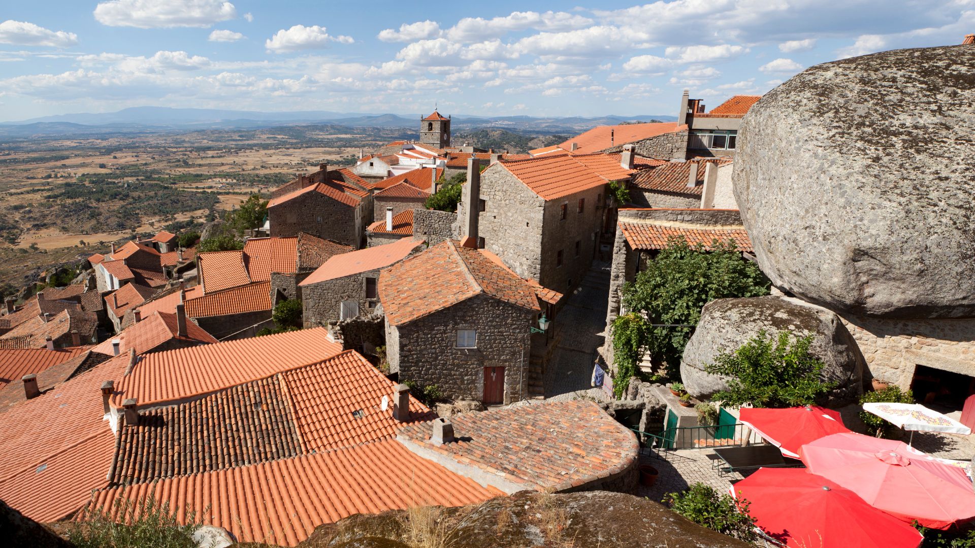 An aerial view showcases the distinctive village of Monsanto in Portugal, characterized by houses built directly into and around massive granite boulders, with red-tiled roofs contrasting against the natural rock formations and a vast landscape extending into the distance under a partly cloudy sky. 