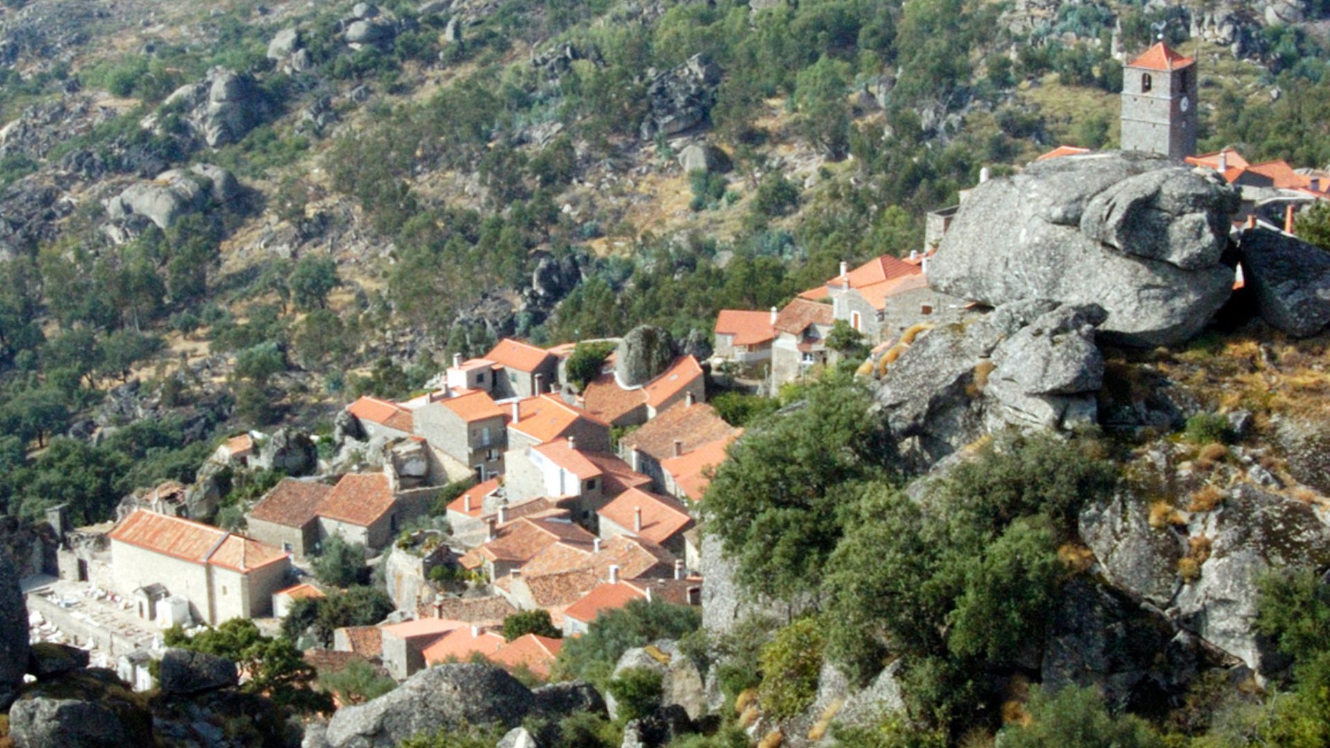 An aerial view showcases the distinctive village of Monsanto in Portugal, characterized by houses built directly into and around massive granite boulders, with red-tiled roofs contrasting against the natural rock formations.