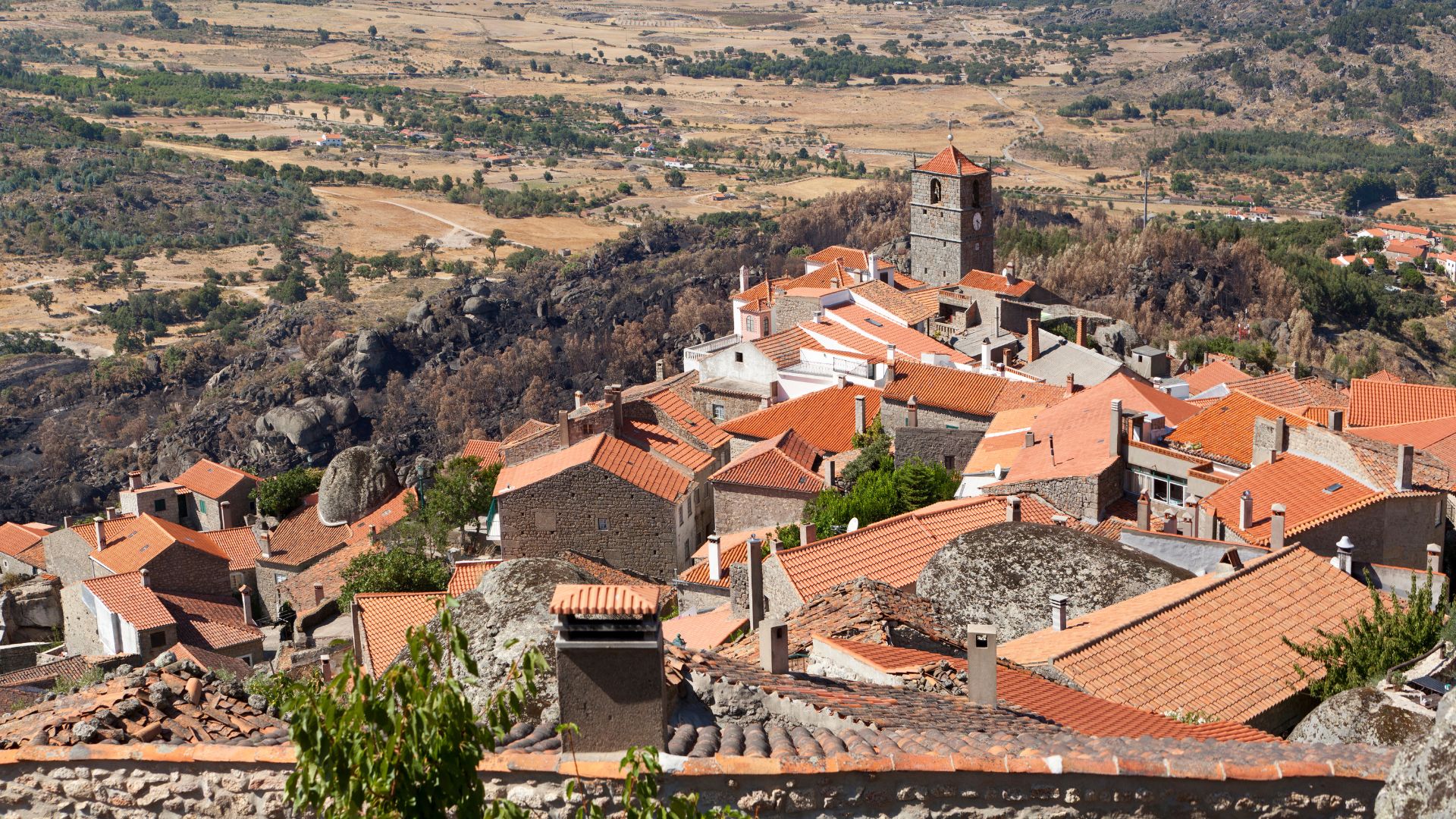 An aerial view showcases the distinctive village of Monsanto in Portugal, characterized by houses built directly into and around massive granite boulders, with red-tiled roofs contrasting against the natural rock formations.
