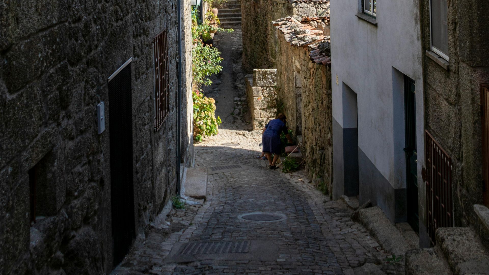 A narrow, cobbled street winds uphill through a historic Monsanto, Central Portugal, flanked by stone buildings and a glimpse of a person tending to plants on the right.