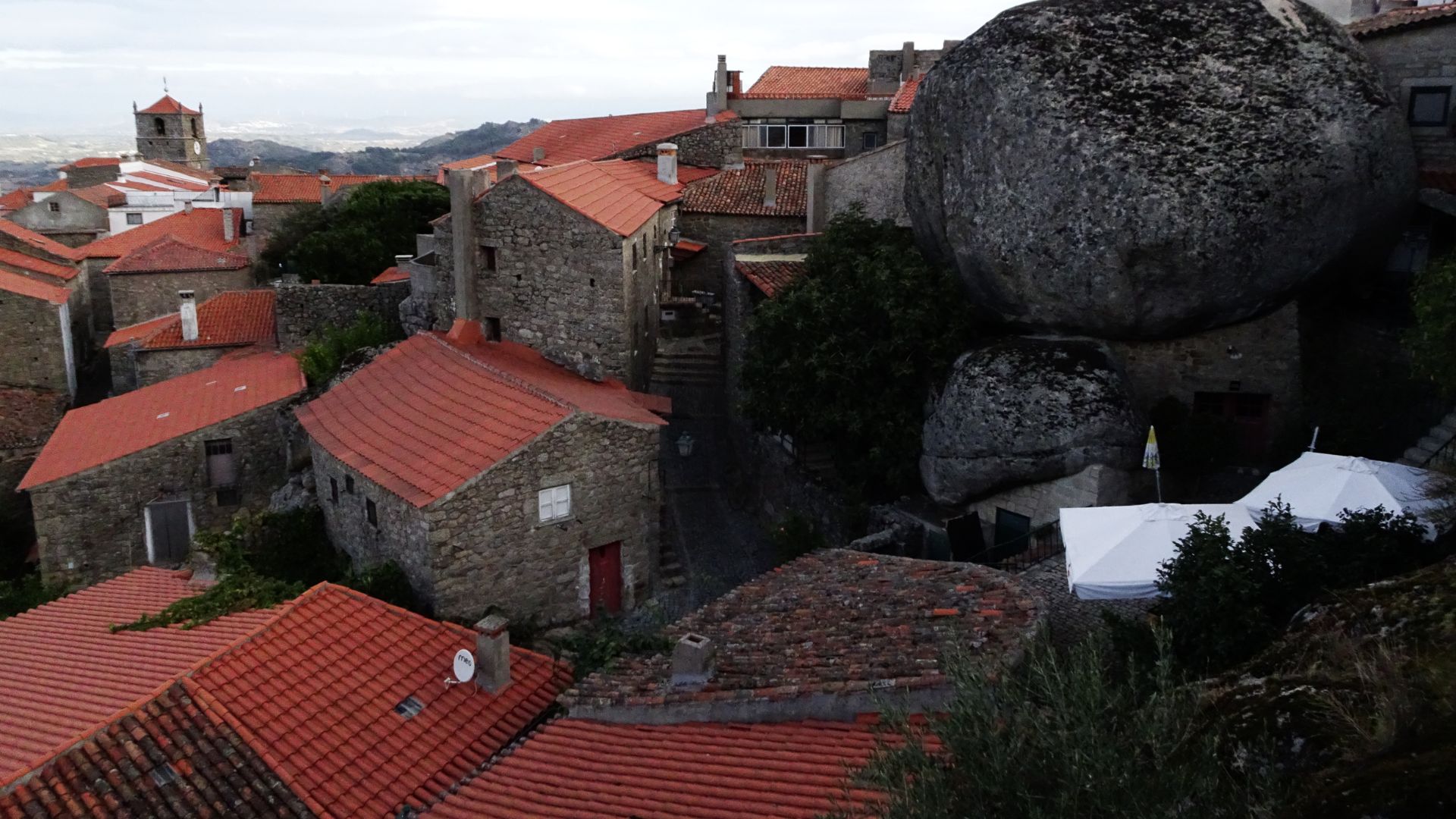 A panoramic view of Monsanto, Portugal, showing traditional stone houses with red-tiled roofs nestled amongst and built into massive granite boulders on a hillside, with a tower visible in the distance.