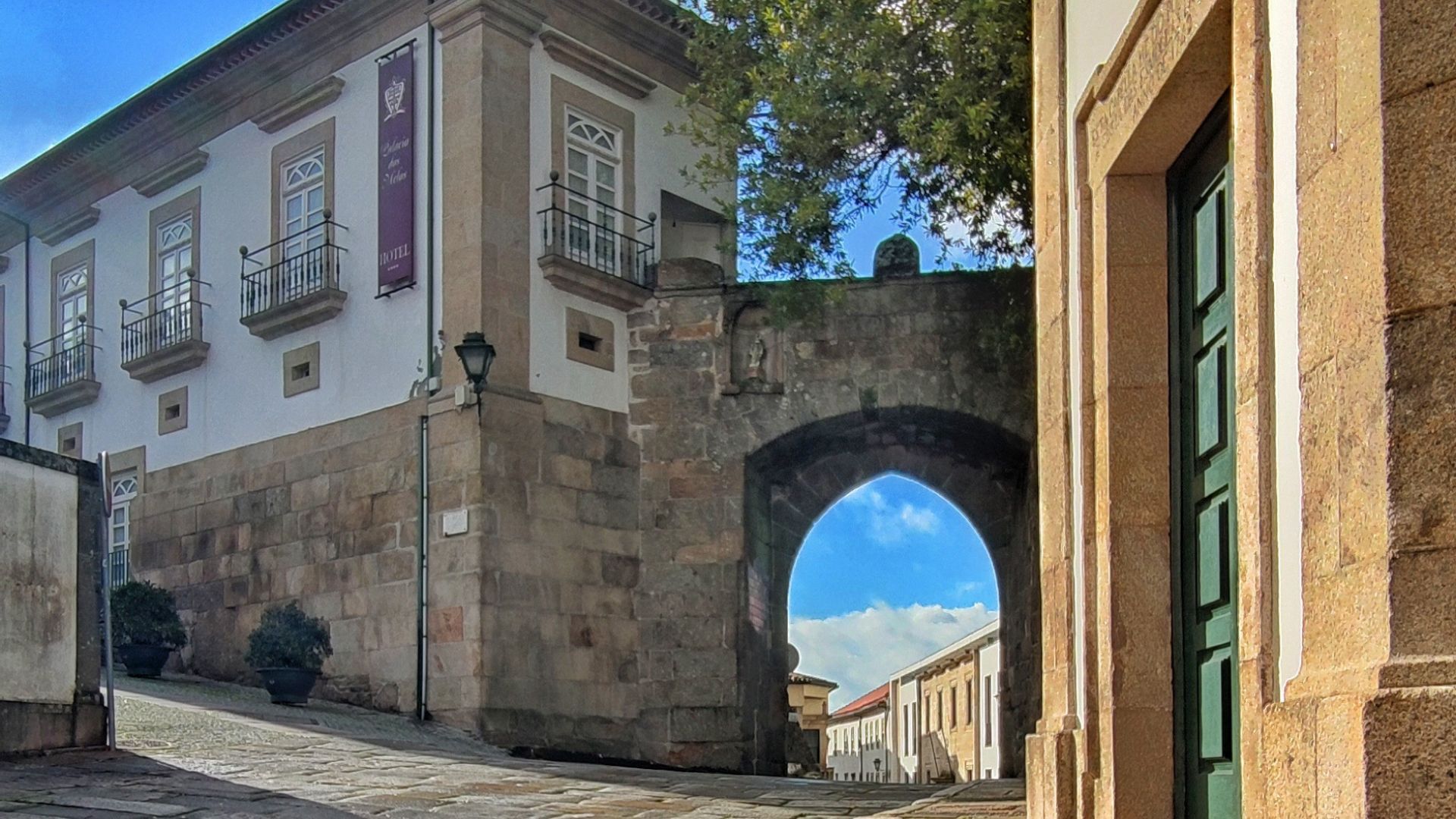 A historic building with a light-colored facade and stone base, featuring balconies with railings, connected to a stone archway that leads to another building with a green door. Blue sky with clouds is visible through the arch.
