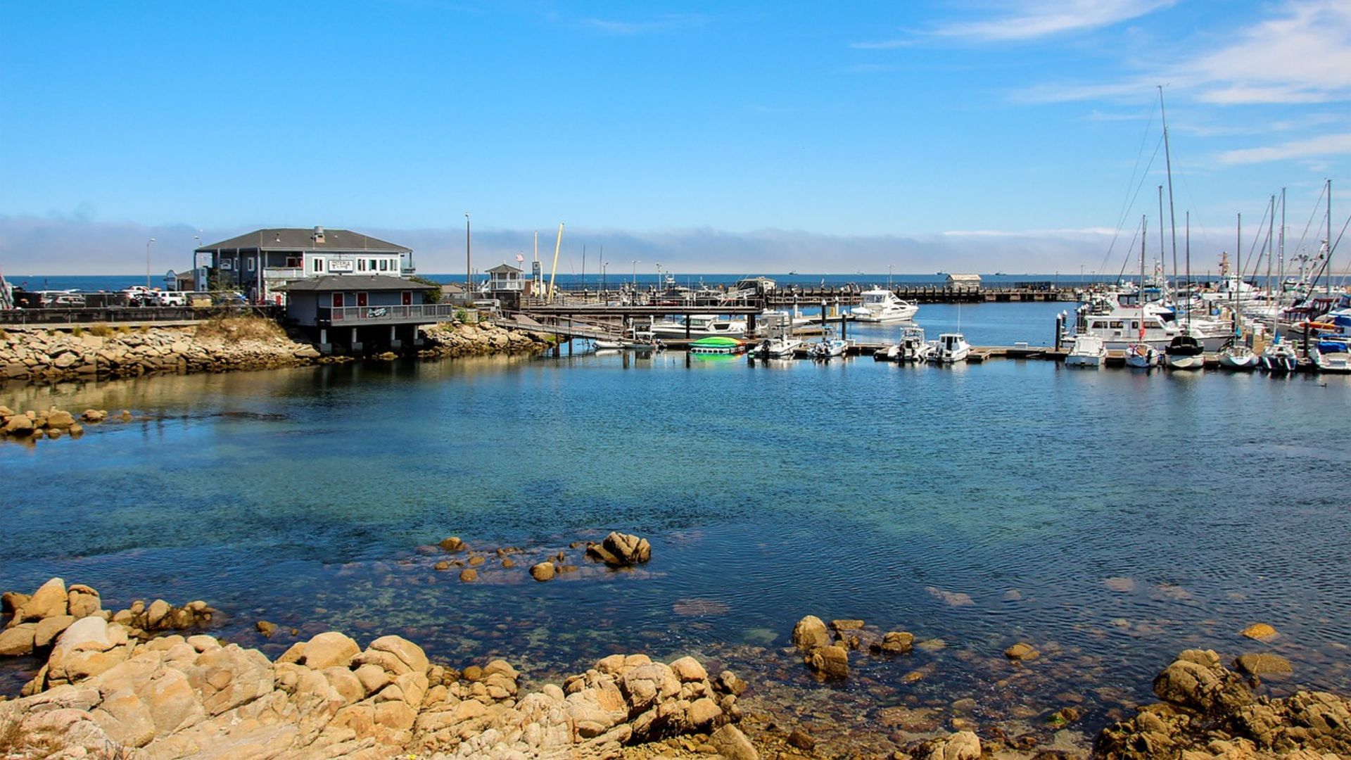 A daytime shot of a calm harbor in Monterey, California, with clear blue water in the foreground, rocky shoreline, and a pier with docked boats and buildings in the background under a bright blue sky.