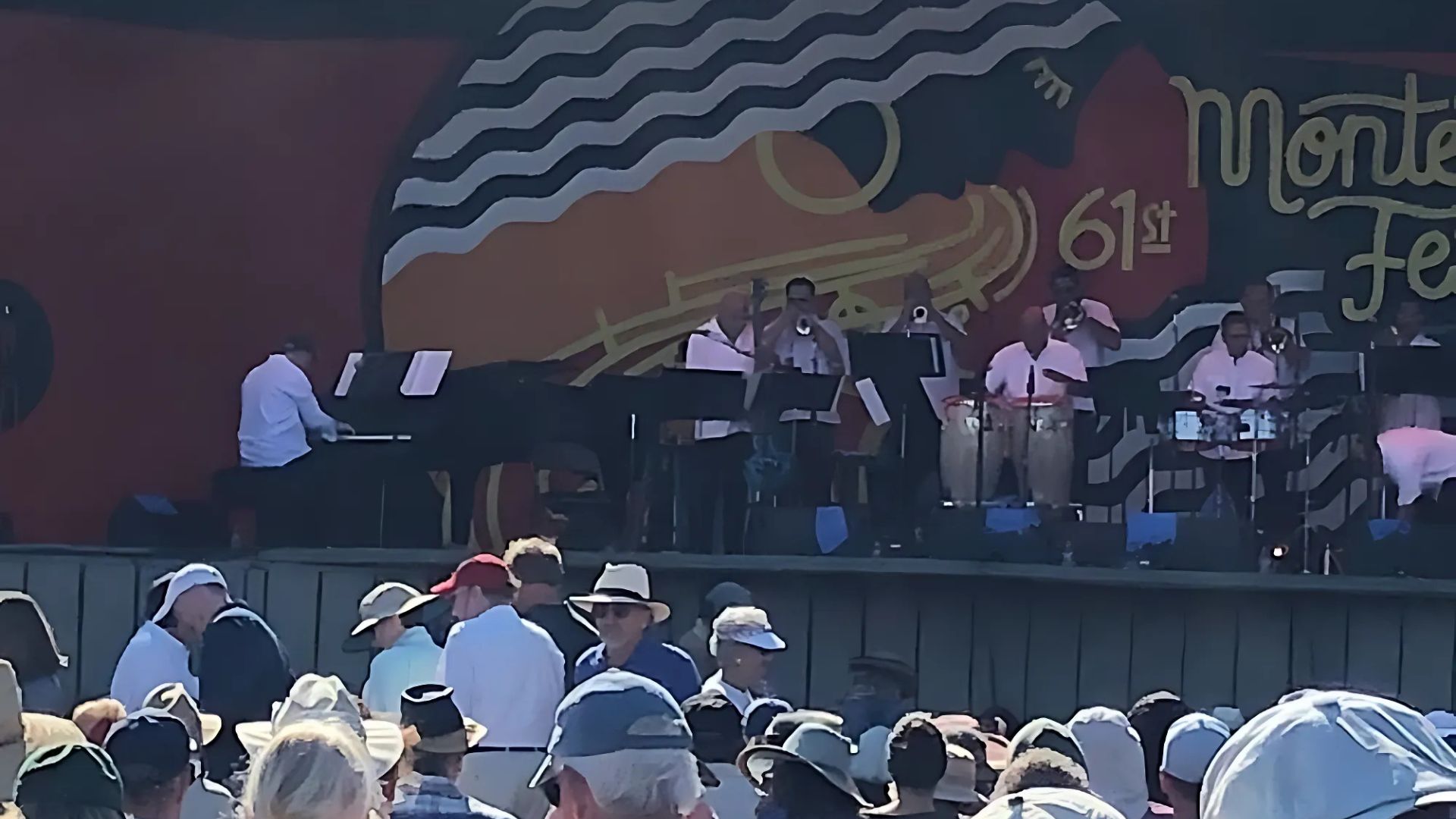 A jazz band performs on stage at the 61st Monterey Jazz Festival, with a large banner displaying the festival name behind them and an audience seated in the foreground.