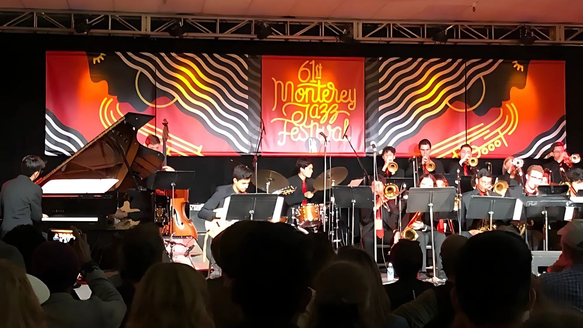 The image captures a live performance at the 61st Monterey Jazz Festival, featuring a jazz ensemble on stage with various instruments including a piano, guitar, drums, and brass instruments, performing in front of an audience. A large red banner with "61st Monterey Jazz Festival" is visible in the background.