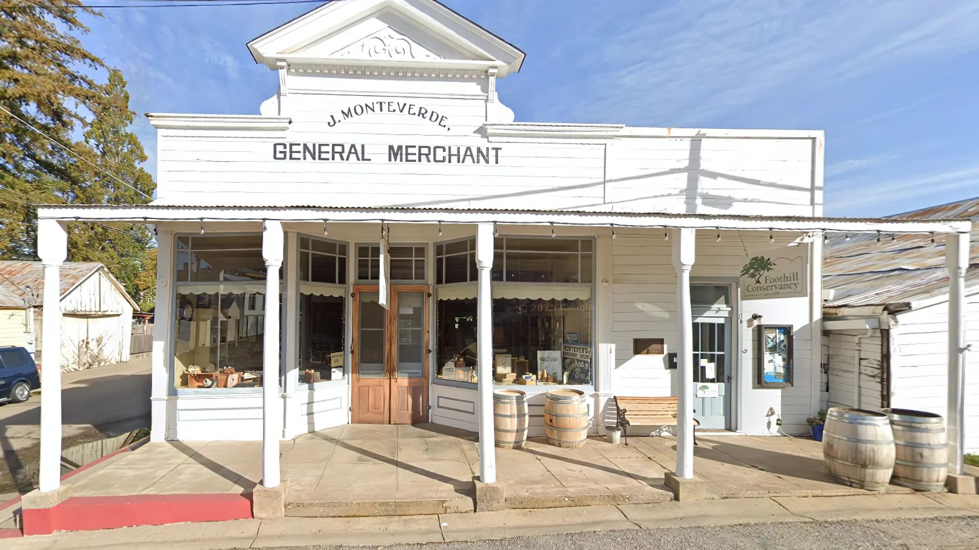 A historic white wooden general store building with a prominent sign reading "J. MONTEVERDE GENERAL MERCHANT" on the facade. The building features large display windows, a welcoming porch with columns, and a double wooden door entrance. Barrels and a wooden bench are placed outside the entrance, and a sign for "Foothill Conservancy" is visible on the right side of the building.