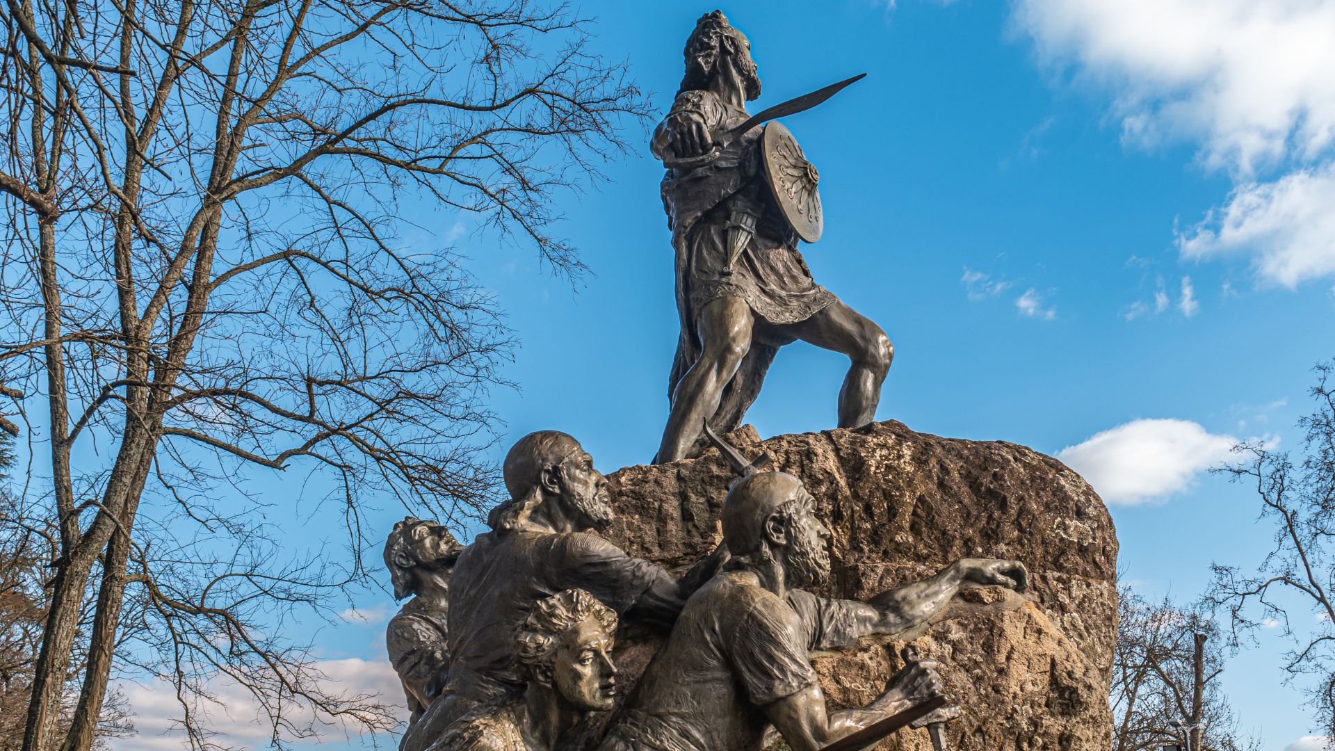 The image shows a bronze statue of Viriathus, a Lusitanian chieftain, standing atop a rock formation, holding a sword and shield. Below him, several other bronze figures are depicted, seemingly in various states of struggle or contemplation. The monument is set against a backdrop of a clear blue sky with some clouds and bare tree branches, suggesting a cooler season. 