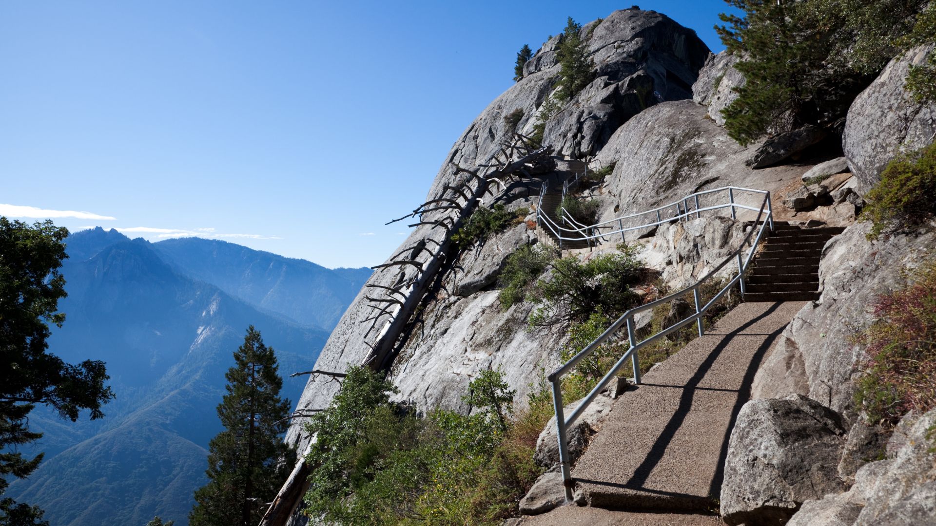 A winding stone and concrete staircase with a metal handrail ascends the side of a large, exposed granite dome, offering panoramic views of mountains and valleys under a clear blue sky.