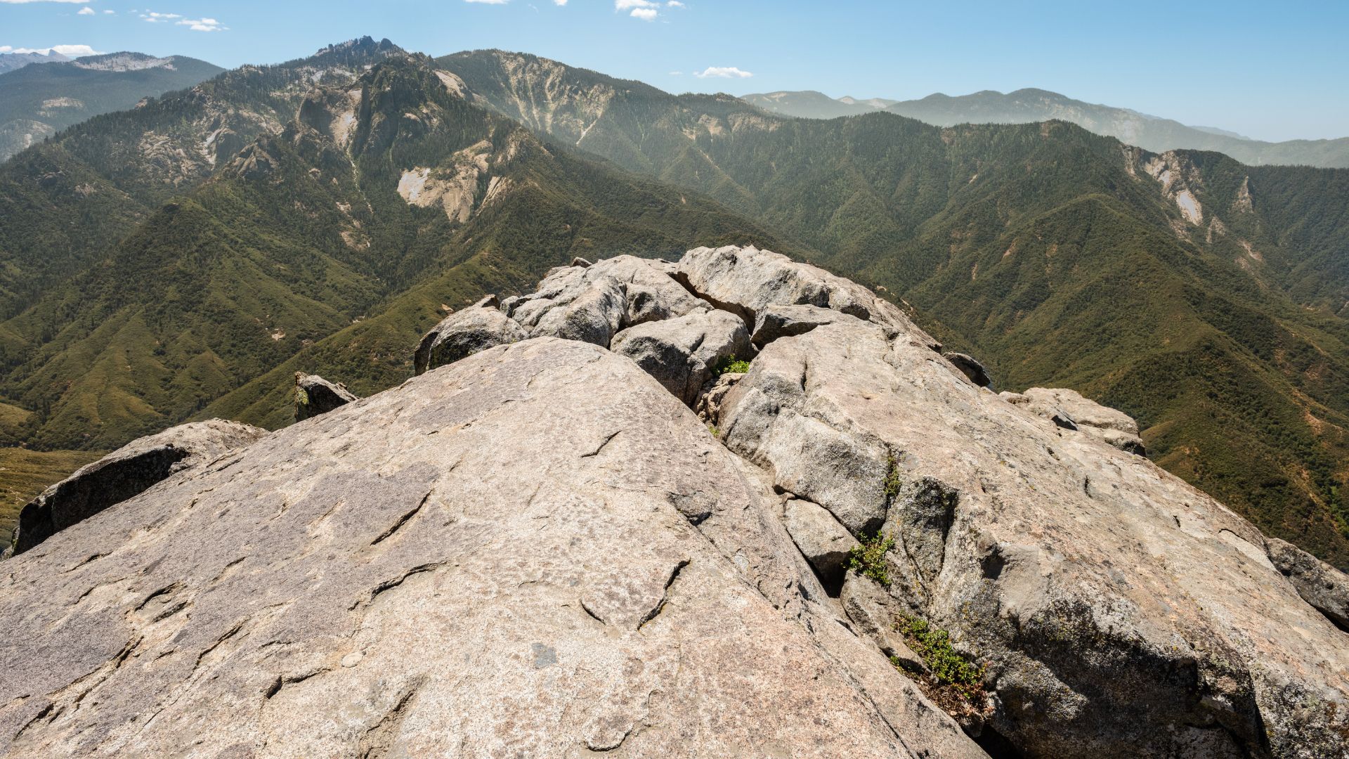 A close-up view from the summit of Moro Rock, a large granite dome, showcasing the rugged rock formations in the foreground and sweeping panoramic views of forested mountains and valleys in the background under a clear sky.