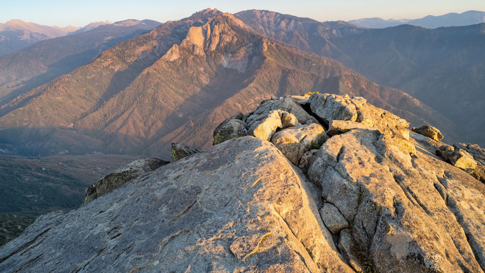 A close-up view of the rugged, sun-drenched granite face of Moro Rock, with the majestic, shadowed peaks of the Great Western Divide visible in the background under a clear sky.