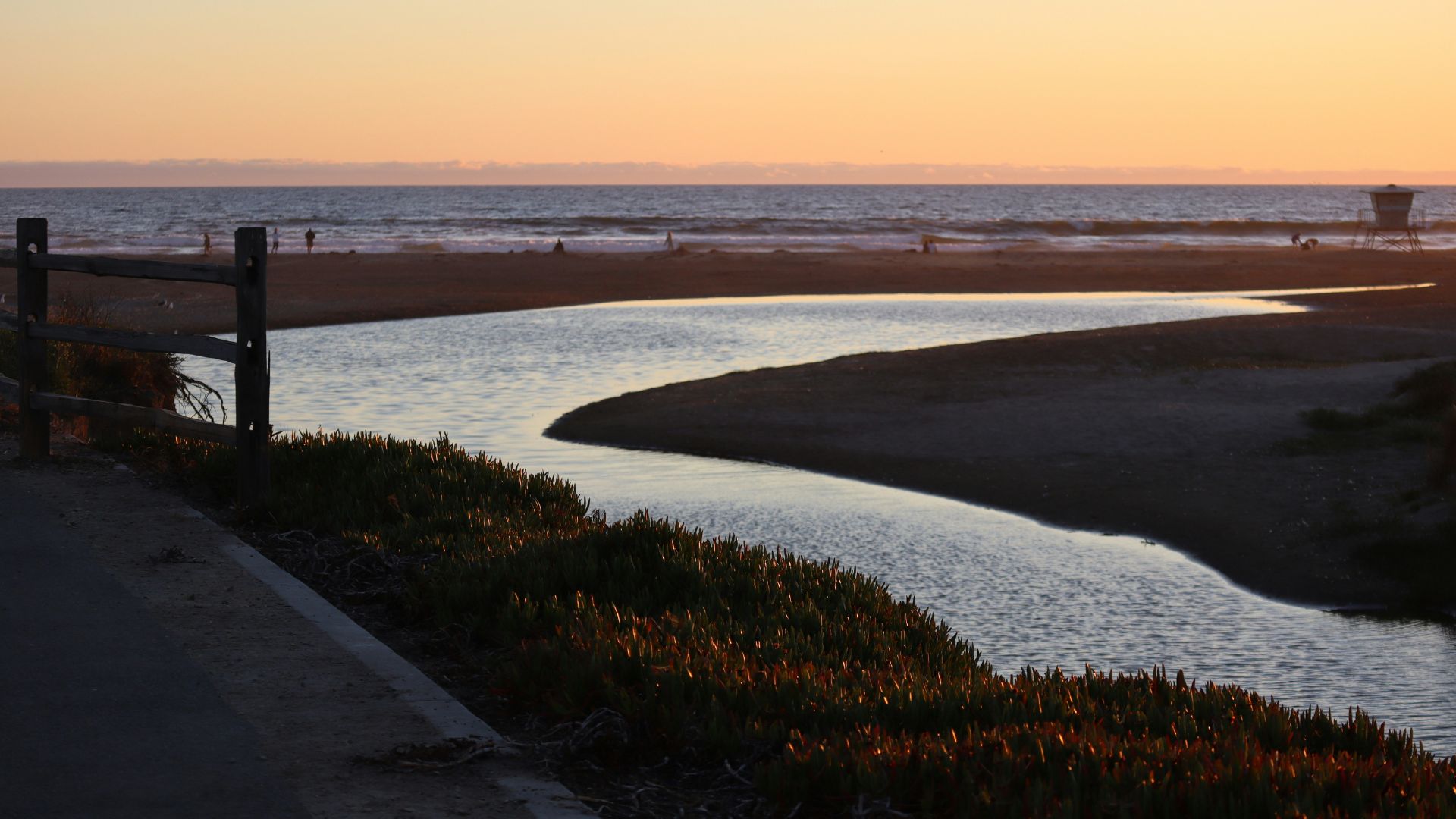 A serene sunset scene at Morro Bay, California, featuring a winding creek flowing into the ocean, with a sandy beach and distant mountains under a warm, orange sky. A wooden fence lines the path on the left, and sparse coastal vegetation is visible in the foreground.
