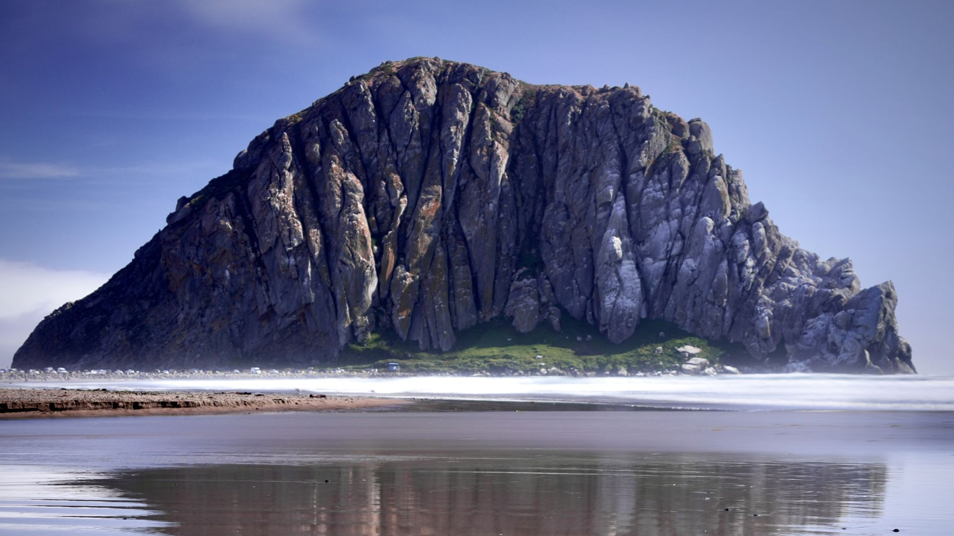 A large, craggy volcanic rock formation, Morro Rock, stands prominently at the edge of the Pacific Ocean, its reflection clearly visible in the calm, wet sand of the beach at low tide, under a bright sky.
