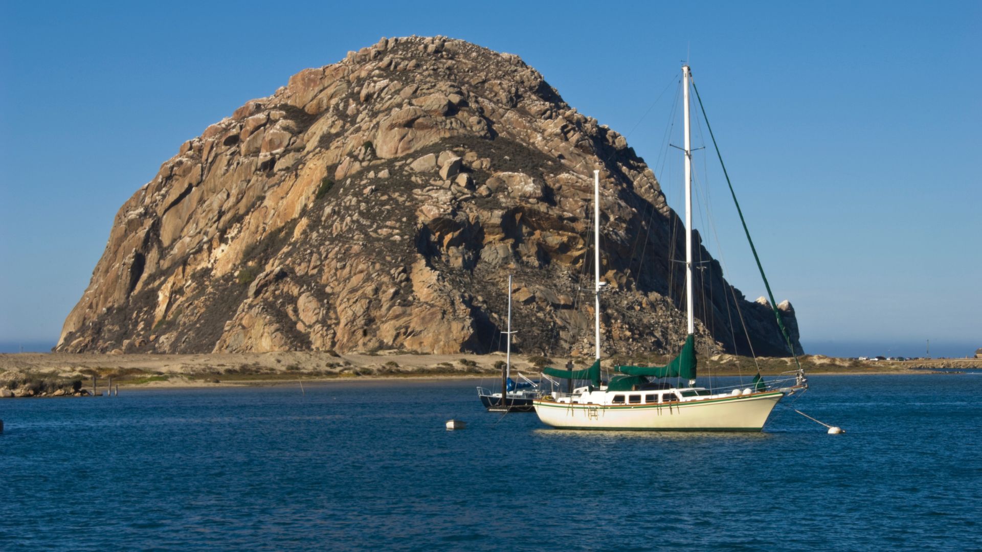 The image captures Morro Rock, a large volcanic plug rising from the sea, with sailboats anchored in the foreground of the calm blue waters of Morro Bay on a clear day. 