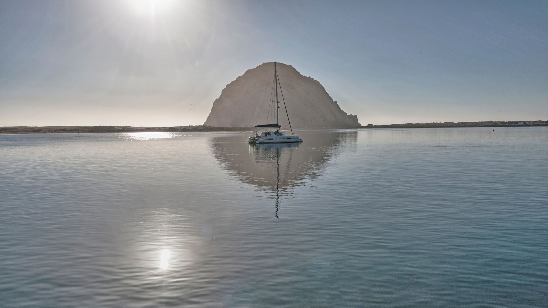 A serene image capturing a sailboat anchored in calm waters with Morro Rock, a large volcanic plug, dominating the background under a bright, sunny sky. The rock's reflection is clearly visible in the tranquil water.