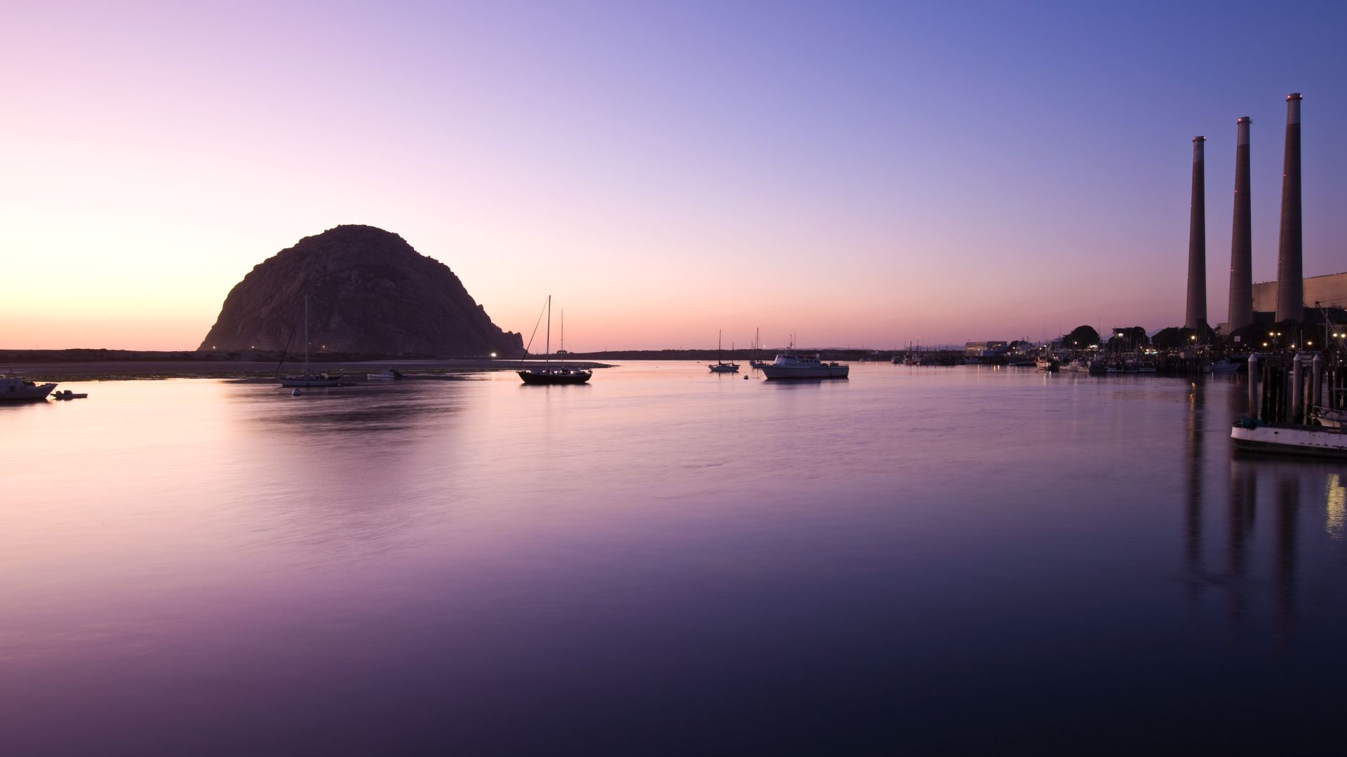 A serene, wide-angle shot of Morro Bay at dusk, showcasing the iconic Morro Rock on the left, boats anchored in the calm water, and the three towering stacks of the Morro Bay Power Plant on the right, all under a gradient sky of purple and orange hues.