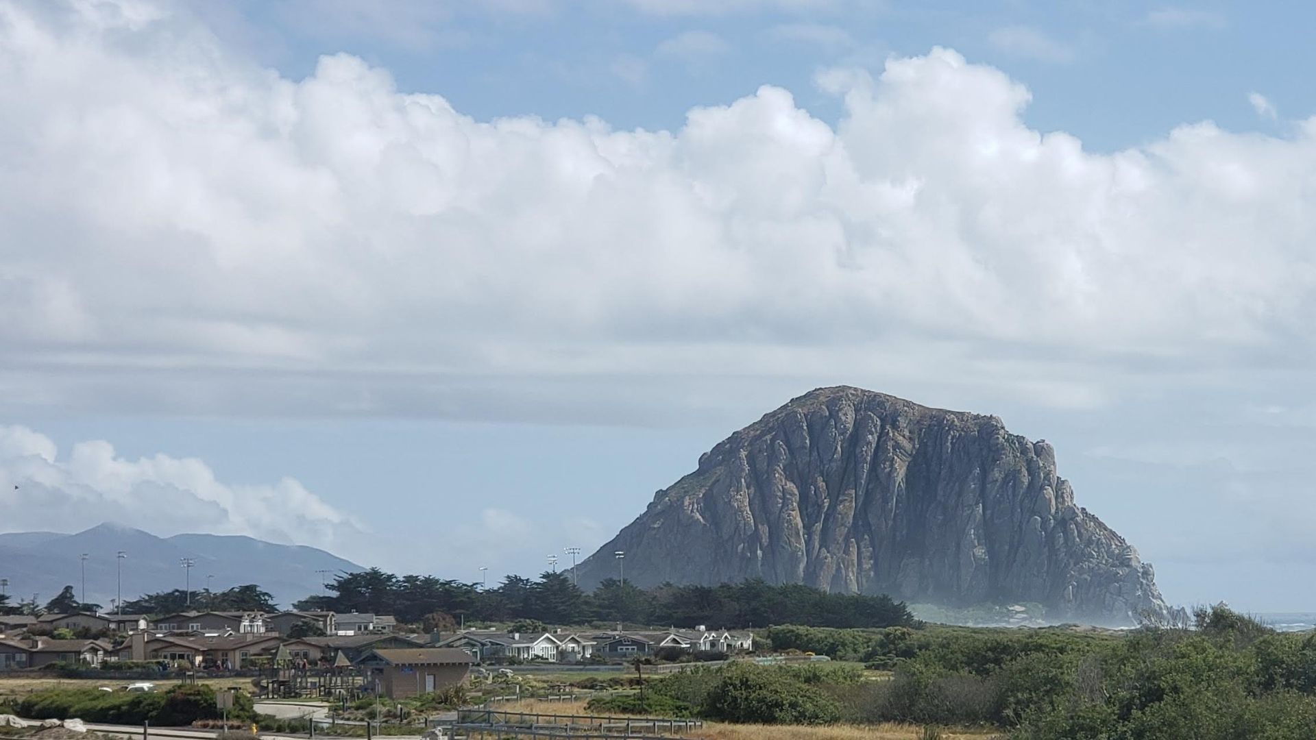 A wide shot of Morro Rock, a large volcanic plug, rising from the Pacific Ocean at the mouth of Morro Bay, with coastal homes and vegetation in the foreground and a cloudy sky above.