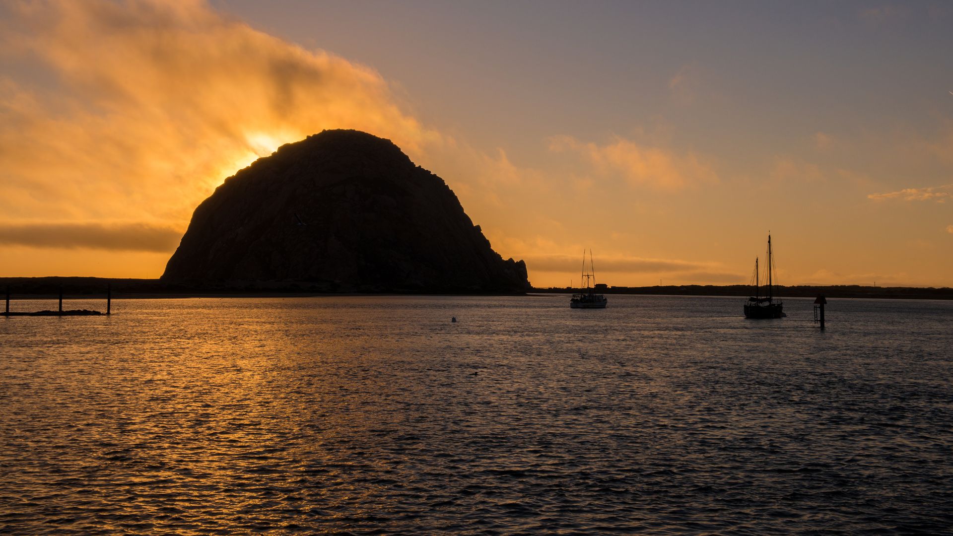 A silhouette of Morro Rock stands prominently against a vibrant orange and yellow sunset over the water of Morro Bay, with two boats visible on the calm water and clouds in the sky.