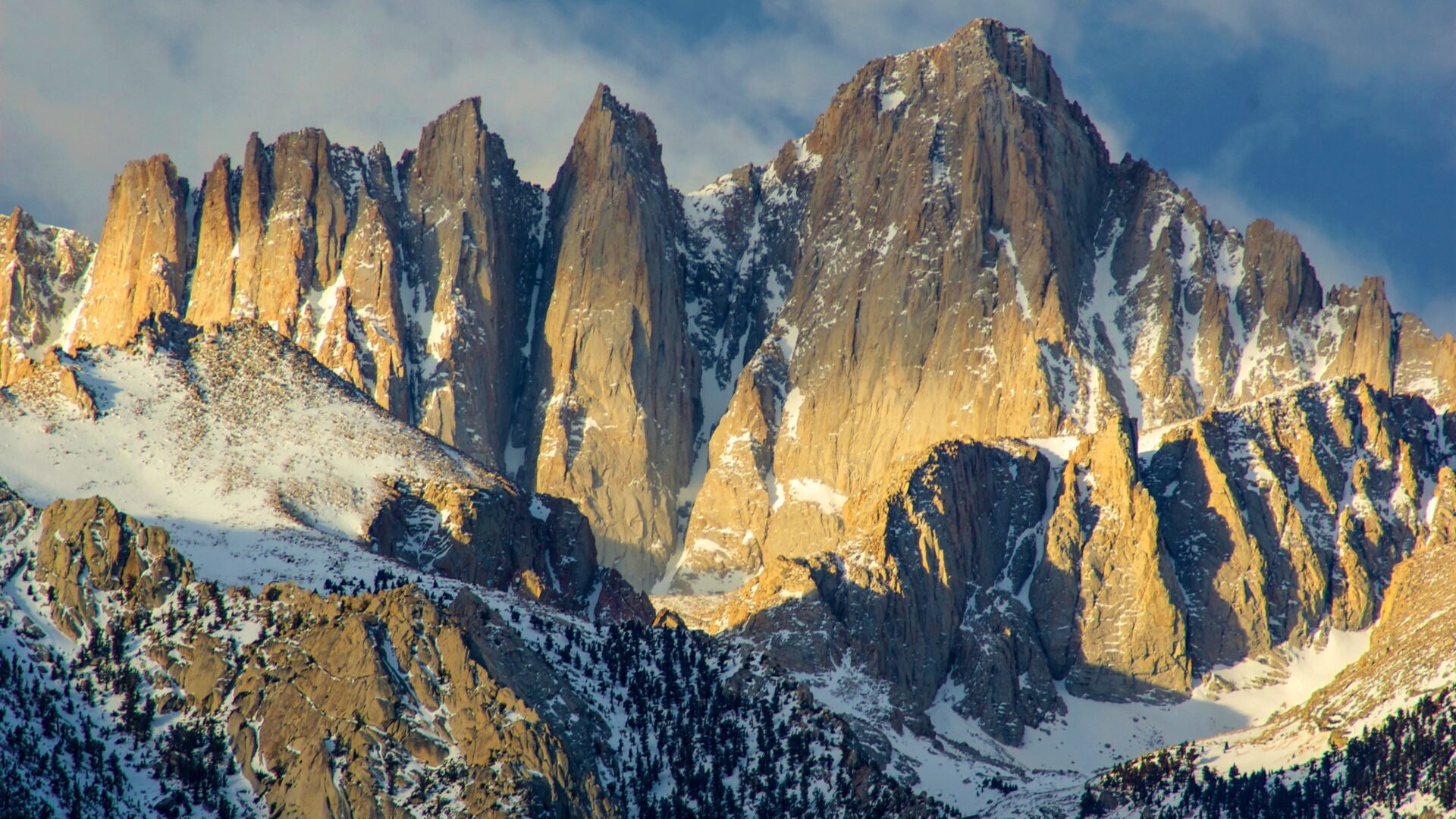 A panoramic view of Mount Whitney in the Sierra Nevada range, with its rugged, snow-dusted peaks illuminated by the warm light of sunrise, and evergreen trees visible in the lower slopes.