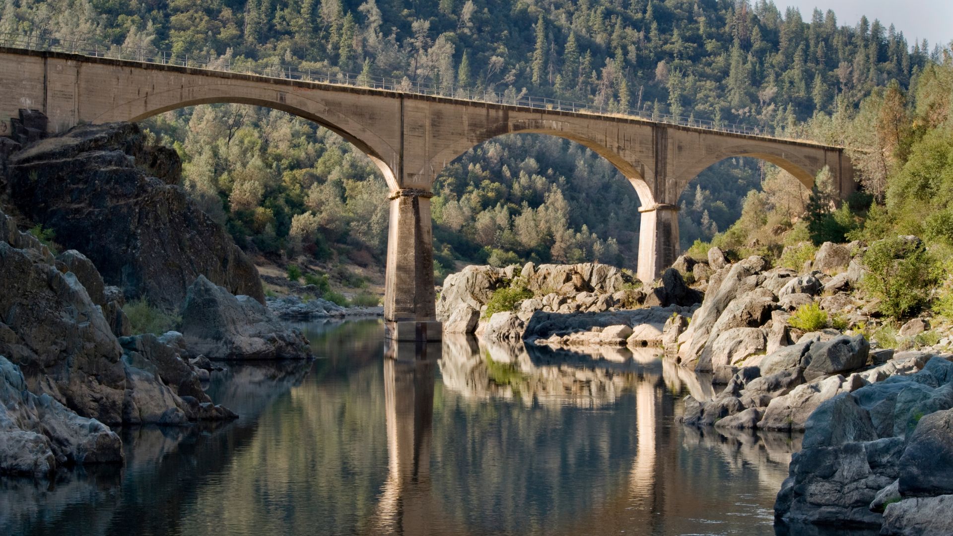 A concrete arch bridge with three prominent arches spans a calm river, reflecting the bridge and the surrounding rocky banks and lush green forested hills under a clear sky.