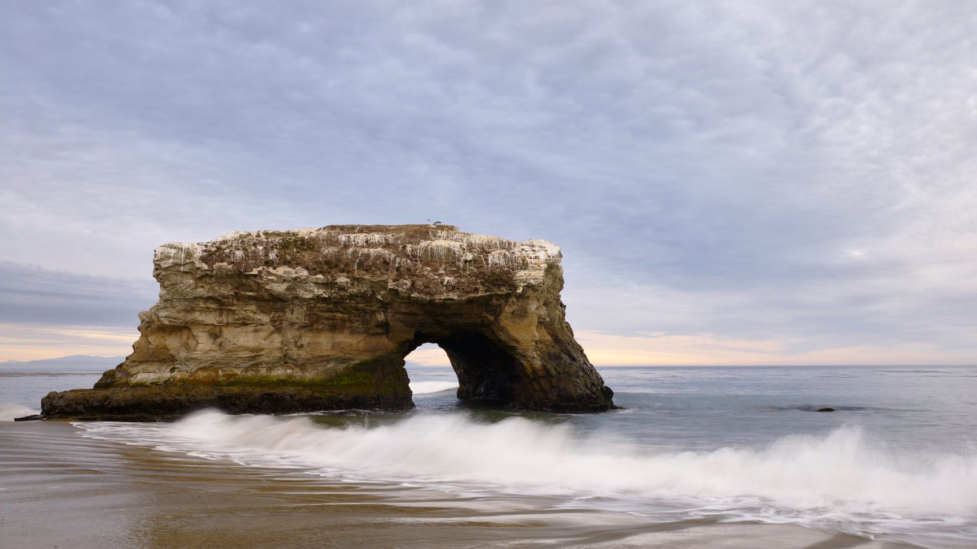 A large natural rock arch formation stands in the ocean at a beach, with waves breaking on the sandy shore under a cloudy sky.