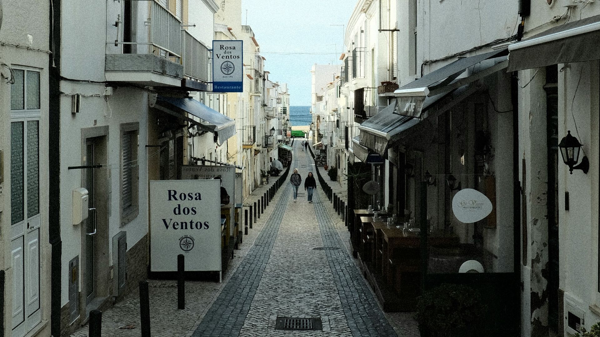 A narrow, cobblestone street lined with white buildings and shops, leading towards the ocean in the distance. Two individuals walk down the street, and signs for "Rosa dos Ventos" are visible on the buildings.