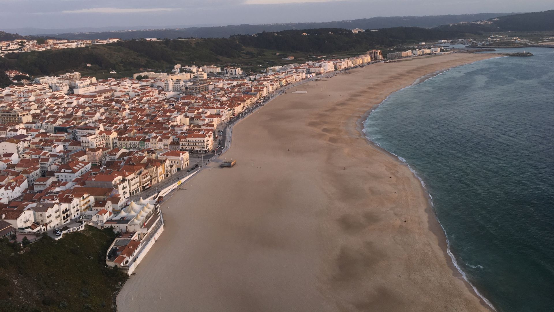 An aerial view showcases the coastal town of Nazaré, Portugal, featuring its expansive sandy beach curving alongside the Atlantic Ocean and a densely built area with white and terracotta-roofed buildings extending inland towards hills. 