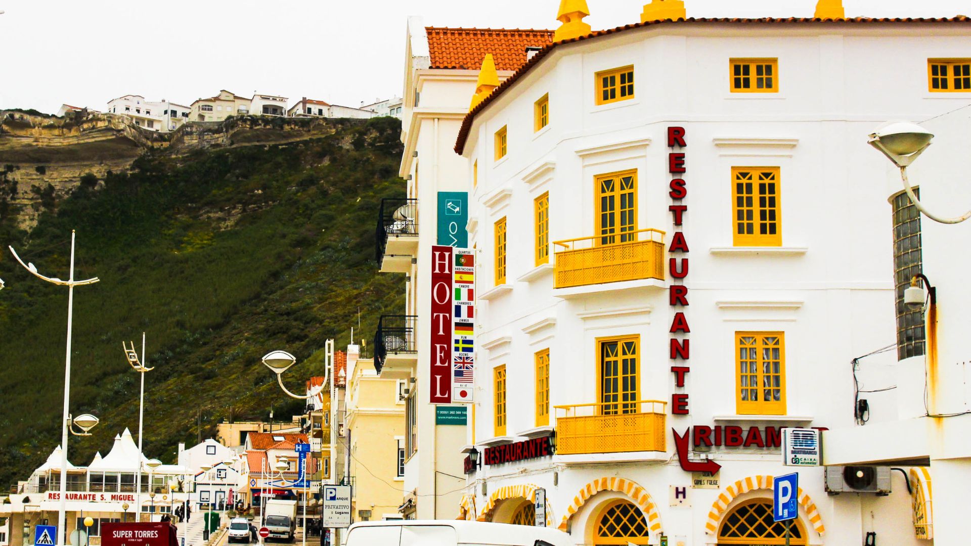 A white multi-story building with yellow window frames and balconies, featuring prominent red signs for "RESTAURANTE RIBAMAR" and a "HOTEL" sign, stands beside a street with lampposts and a hillside in the background. 