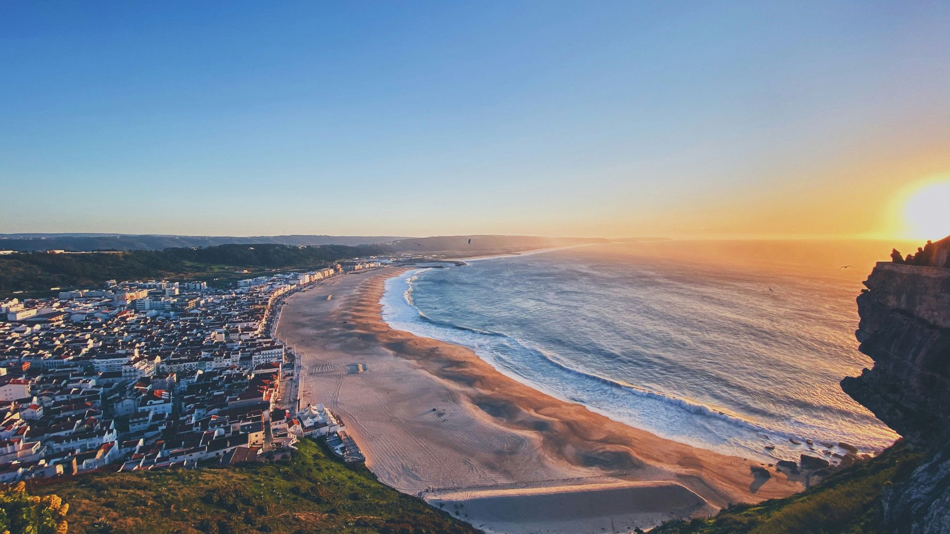 An aerial view captures the scenic coastline of Nazaré, Portugal, featuring a wide sandy beach, the town's buildings, and the vast Atlantic Ocean under a clear sky during sunset. 