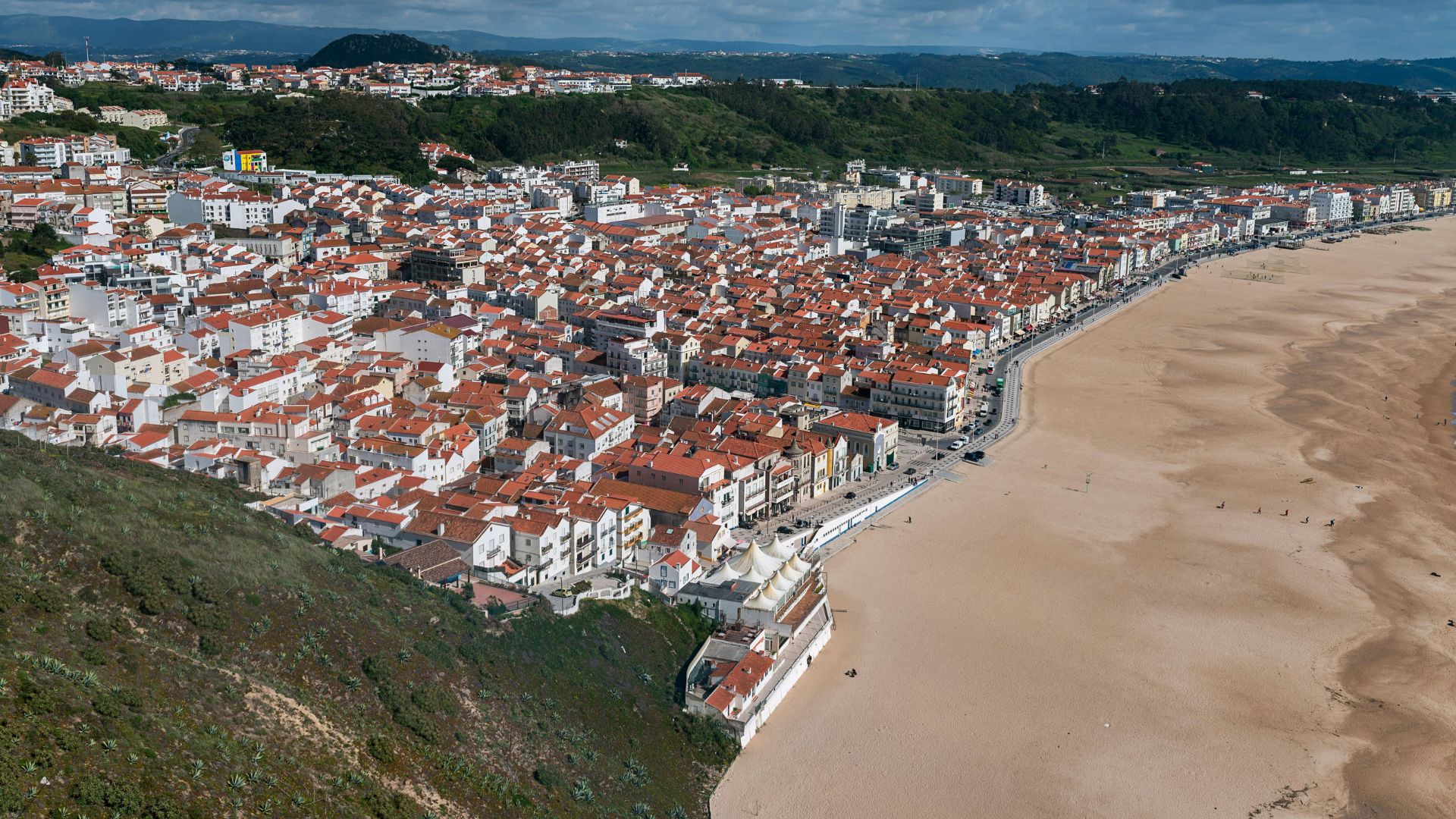  An aerial view of the coastal town of Nazaré, Portugal, showing densely packed white and terracotta-roofed buildings alongside a wide sandy beach and the Atlantic Ocean.