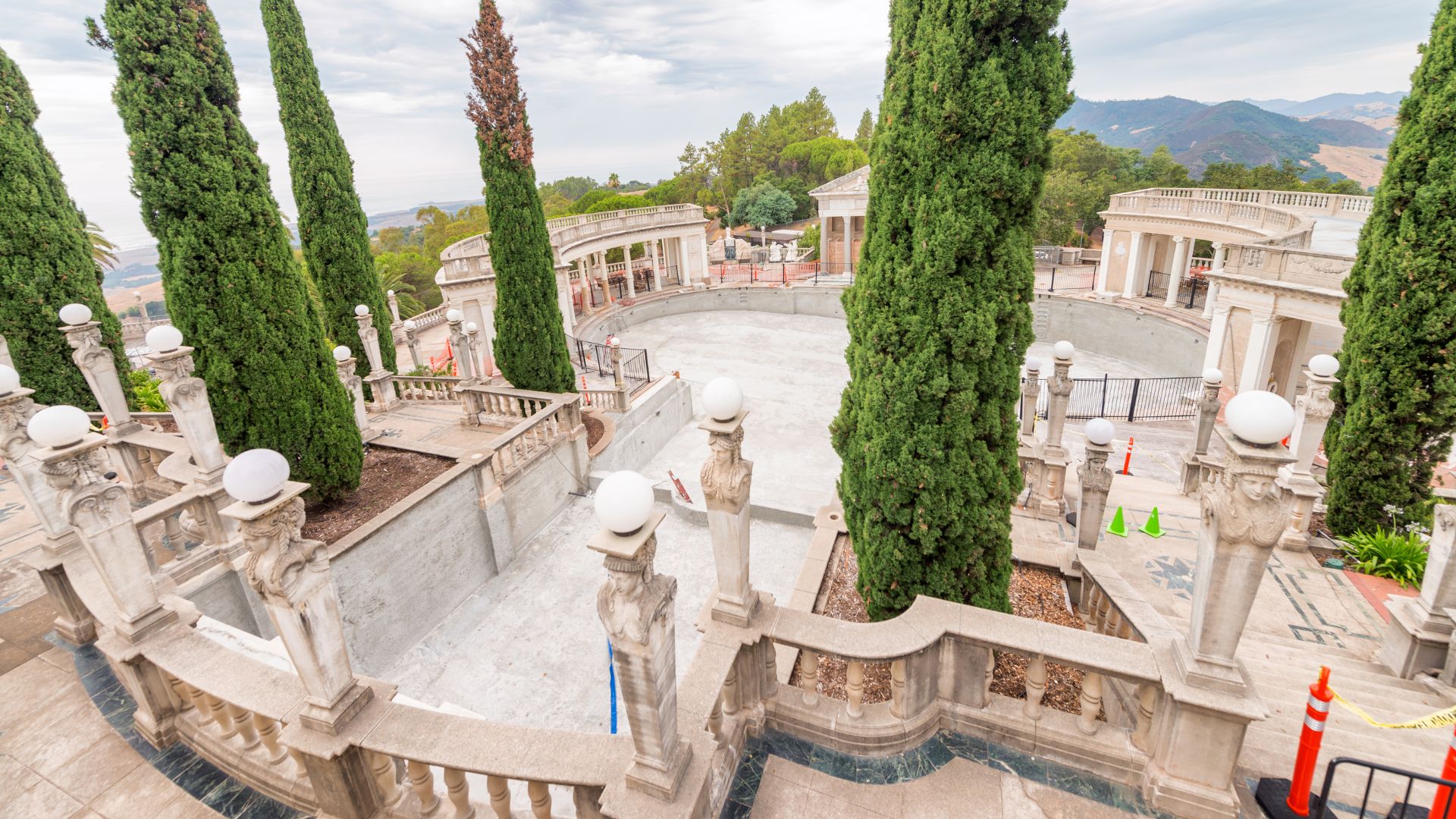An elevated view of the Neptune Pool at Hearst Castle, showcasing its large, ornate outdoor swimming pool surrounded by classical architecture, statues, and tall, slender evergreen trees, with a glimpse of rolling hills and the Pacific Ocean in the distance under a cloudy sky. 