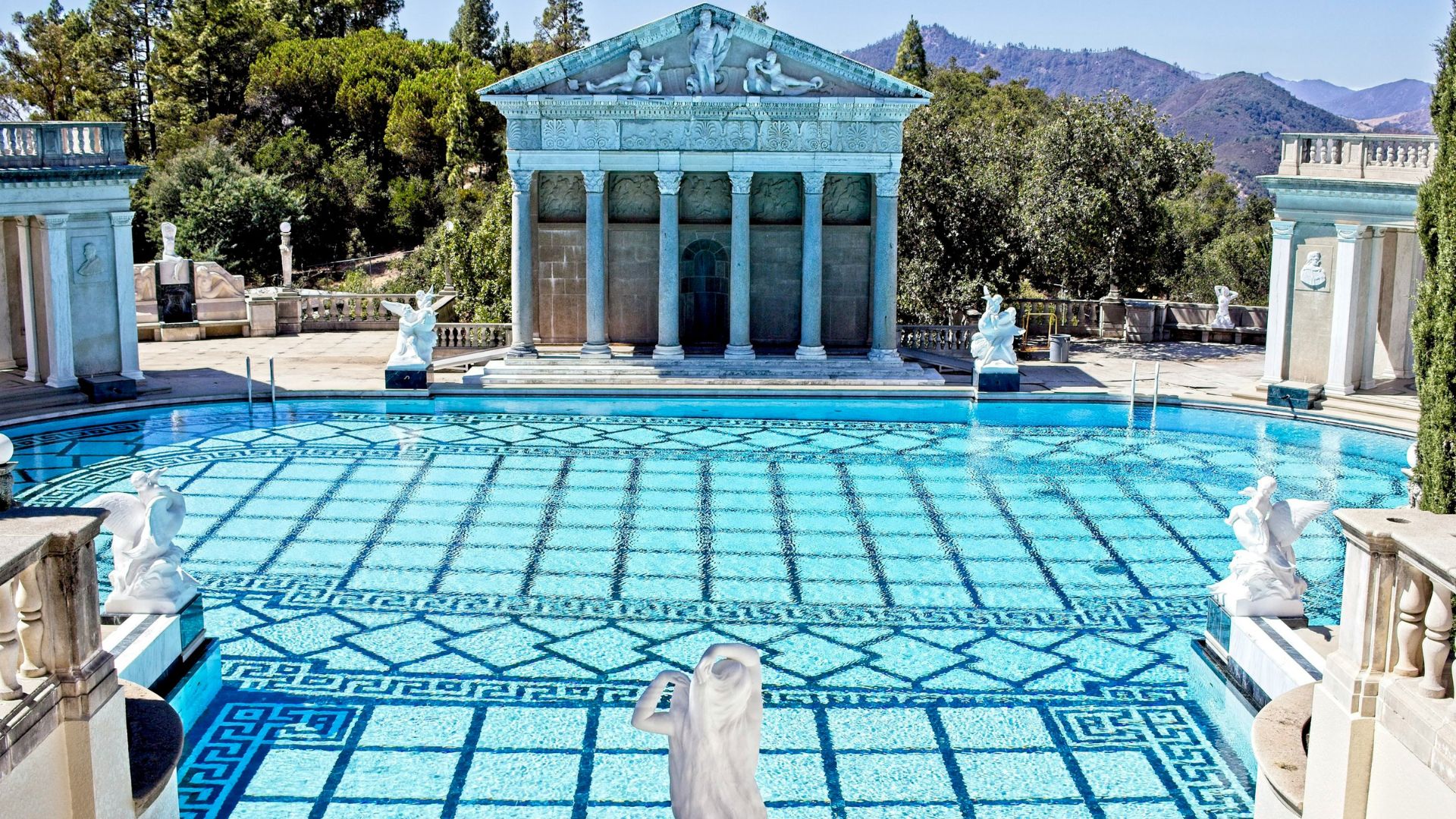 A wide shot captures the iconic Neptune Pool at Hearst Castle, featuring a large, ornate outdoor swimming pool with a temple-like structure at its far end and classical statues surrounding the pool and along its edges, all set against a backdrop of lush green hills and a clear blue sky.