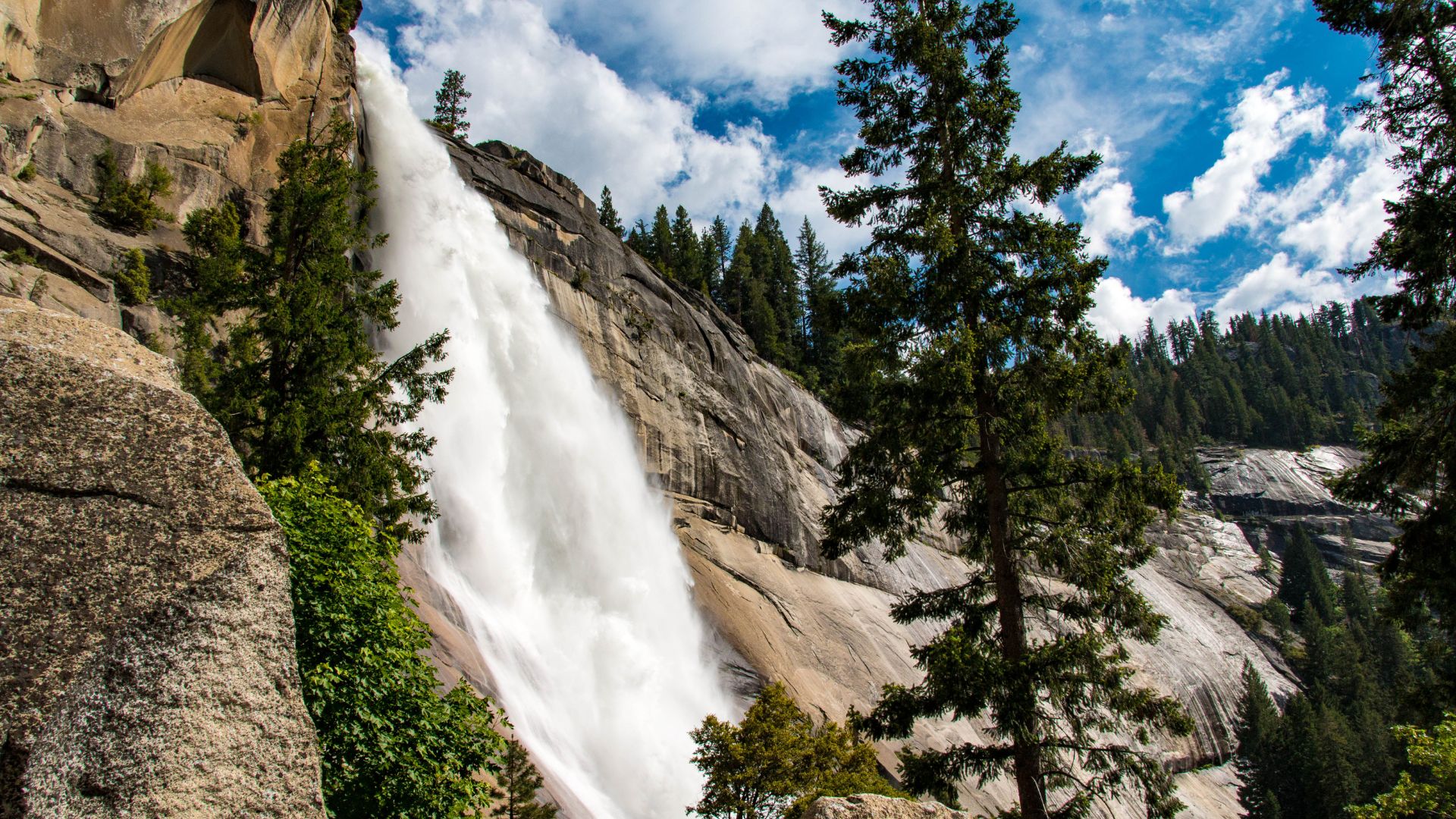A majestic waterfall, Nevada Fall, cascades down a steep granite cliff face surrounded by lush green trees and rocky terrain under a bright blue sky with scattered white clouds in Yosemite National Park, California. 