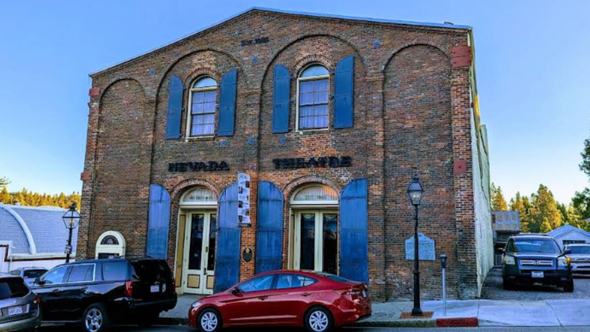 A historic brick building with arched windows and blue shutters, labeled "NEVADA THEATRE," stands on a street with parked cars in front of it under a clear blue sky.