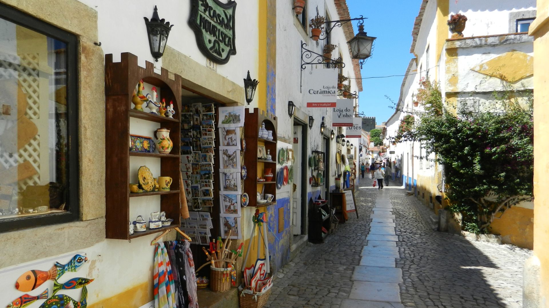 The image shows a narrow, cobblestone street lined with whitewashed buildings and shops in Óbidos, Portugal. On the left, a shop with colorful pottery and textiles displayed outside is visible, while the right side features more buildings and a lush green bush, leading down the winding street under a clear blue sky. 