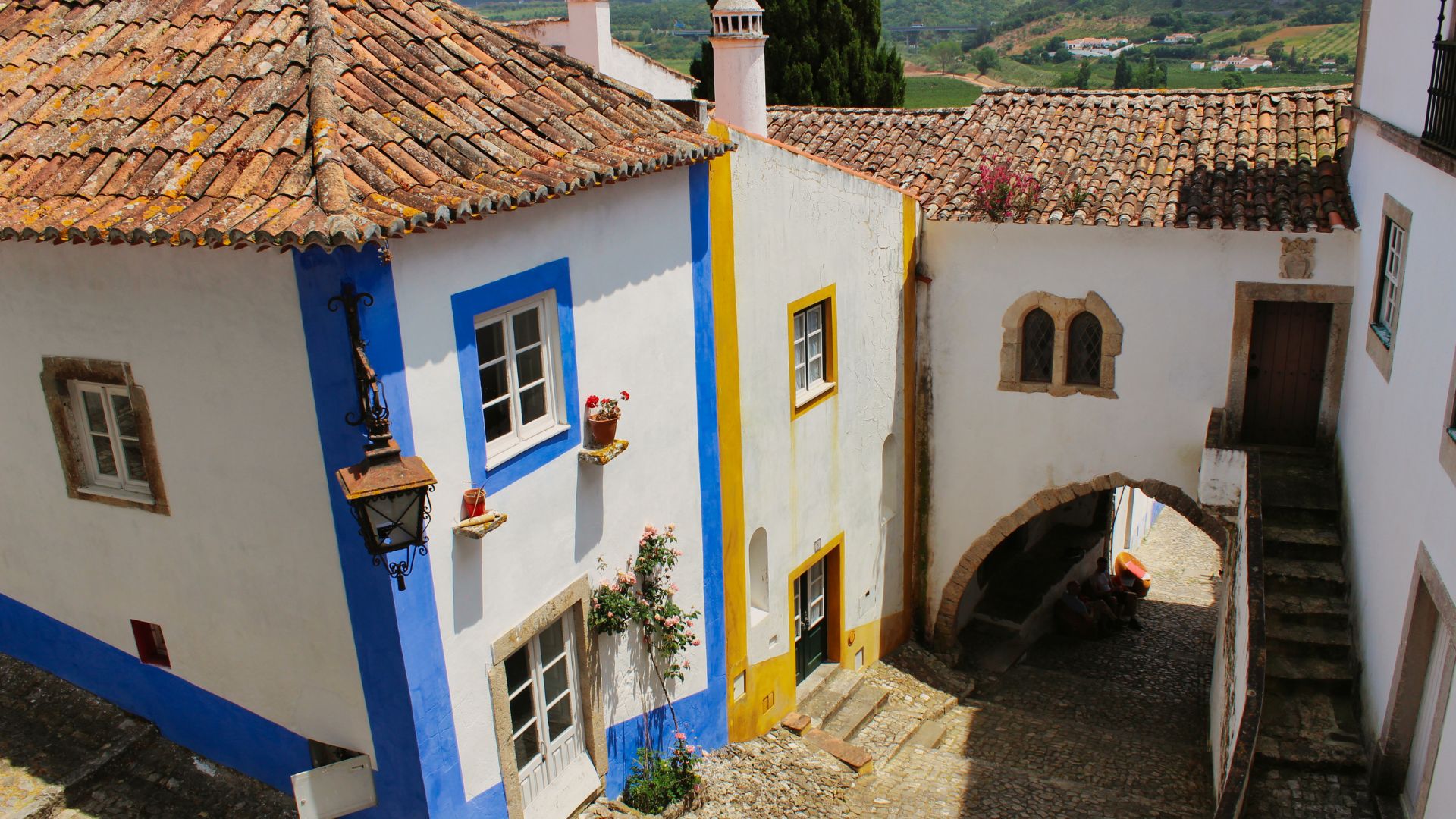 A sunlit view of the Arco da Cadeia in Óbidos, Portugal, featuring whitewashed buildings with blue and yellow accents, terracotta tiled roofs, and a prominent arched passageway leading to a cobblestone courtyard.