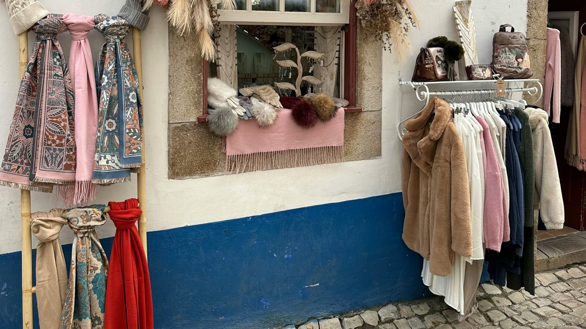 A street scene featuring a shop's outdoor display of colorful scarves, hats, and coats against a white building with a blue base and a window adorned with dried foliage.