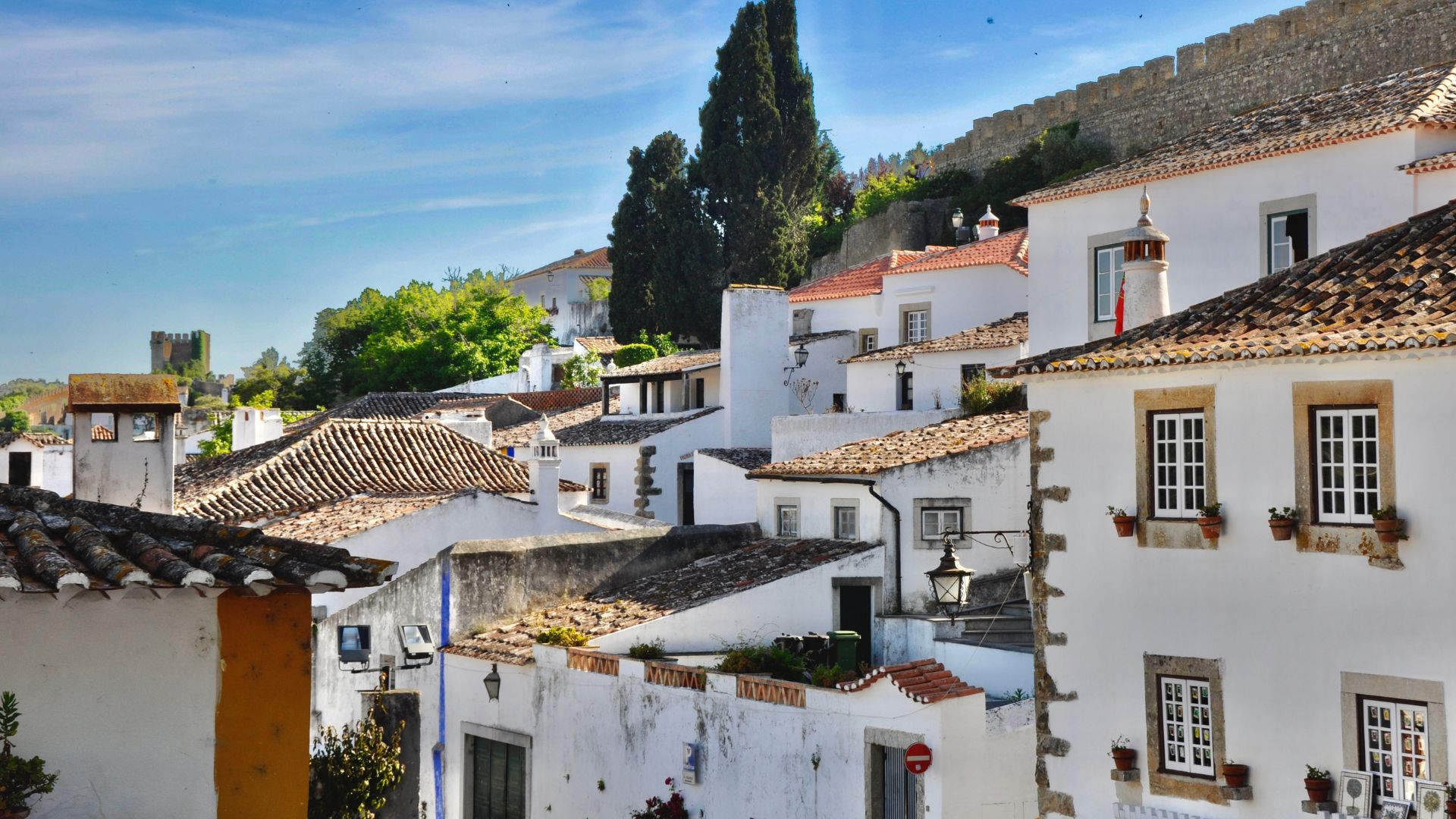 A picturesque view of the medieval town of Óbidos, Portugal, featuring whitewashed houses with terracotta roofs and a castle wall on a hill under a blue sky.