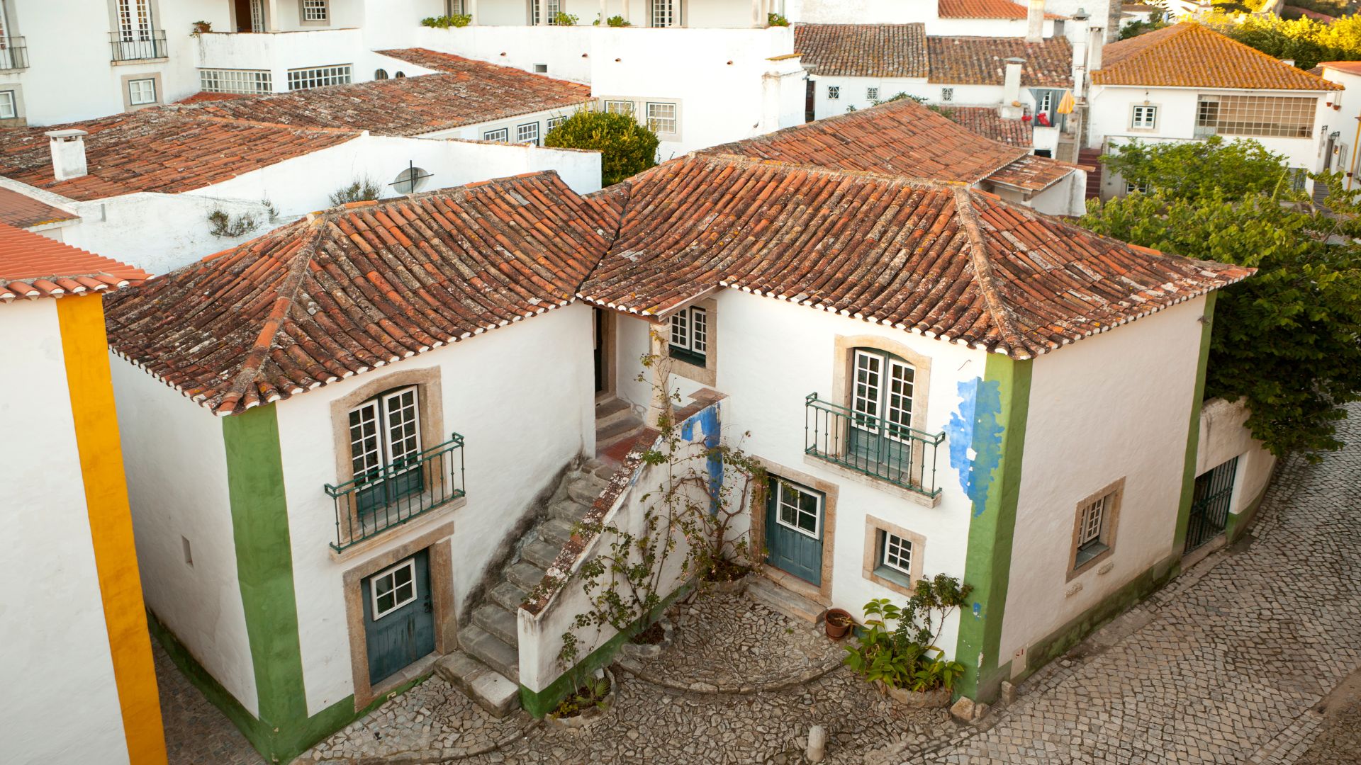 The image shows a charming, high-angle view of whitewashed houses with traditional terracotta tiled roofs and green and blue accents in the historic, walled town of Óbidos, Portugal. The cobblestone street and lush greenery add to the medieval atmosphere. 