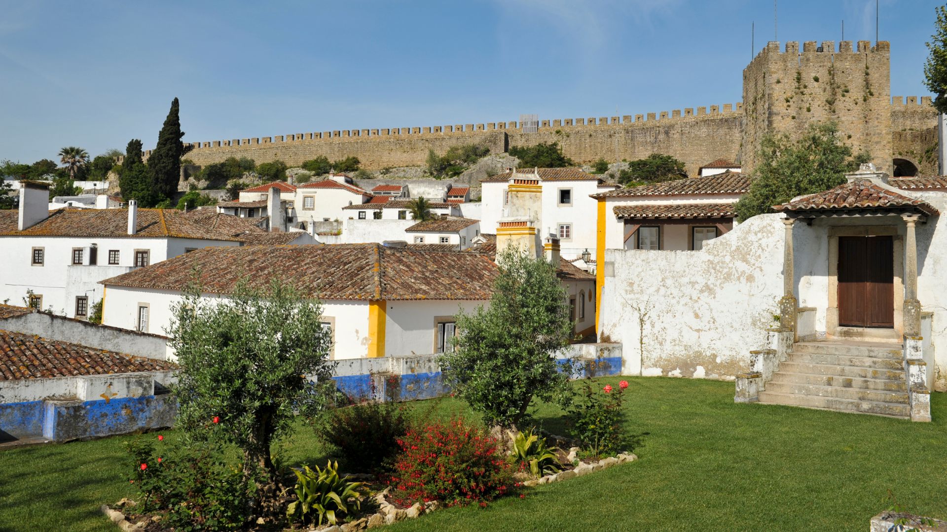 A panoramic view of the medieval walled town of Óbidos, Portugal, featuring whitewashed buildings with terracotta roofs in the foreground and the imposing stone walls and towers of the Castelo de Óbidos on a hilltop in the background under a clear blue sky.