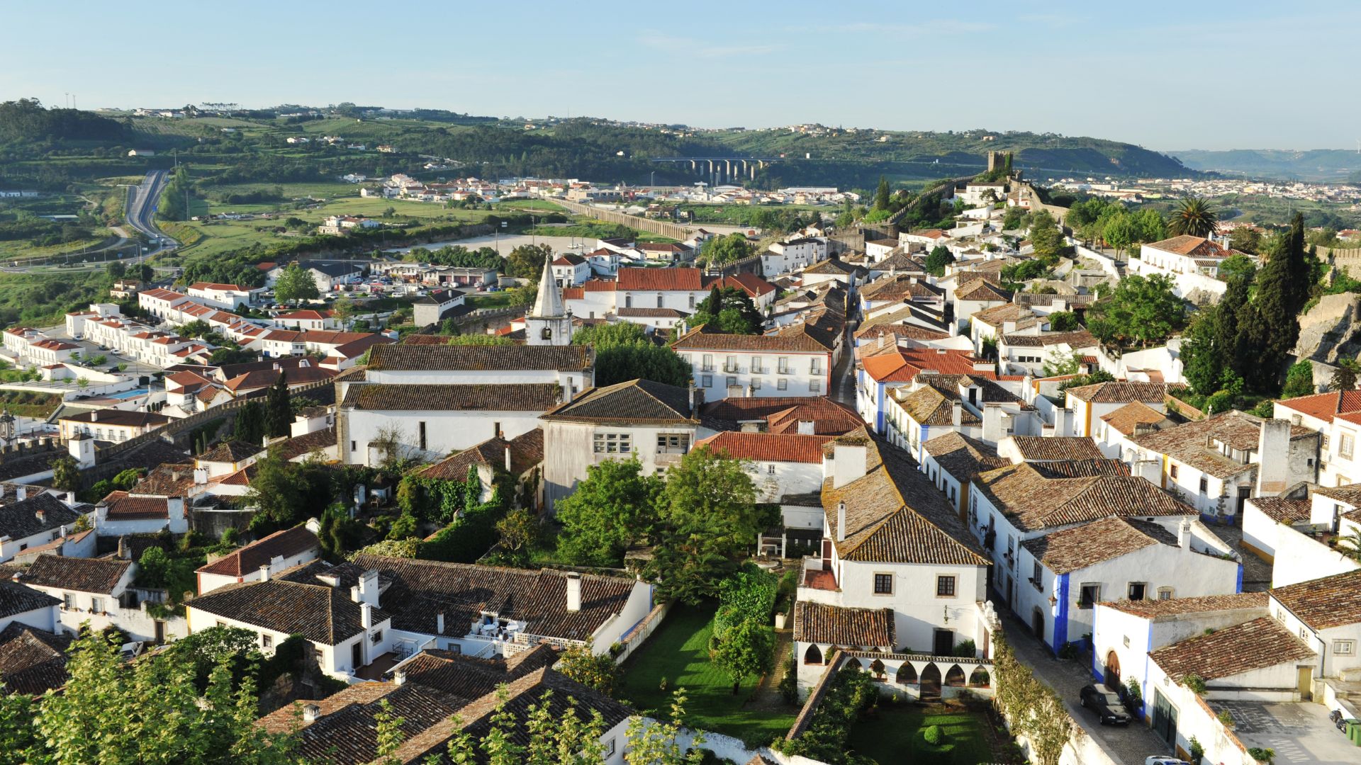 A panoramic view of the charming, walled medieval town of Óbidos, Portugal, with its white-washed buildings and terracotta roofs nestled amidst green hills, leading up to a historic castle.