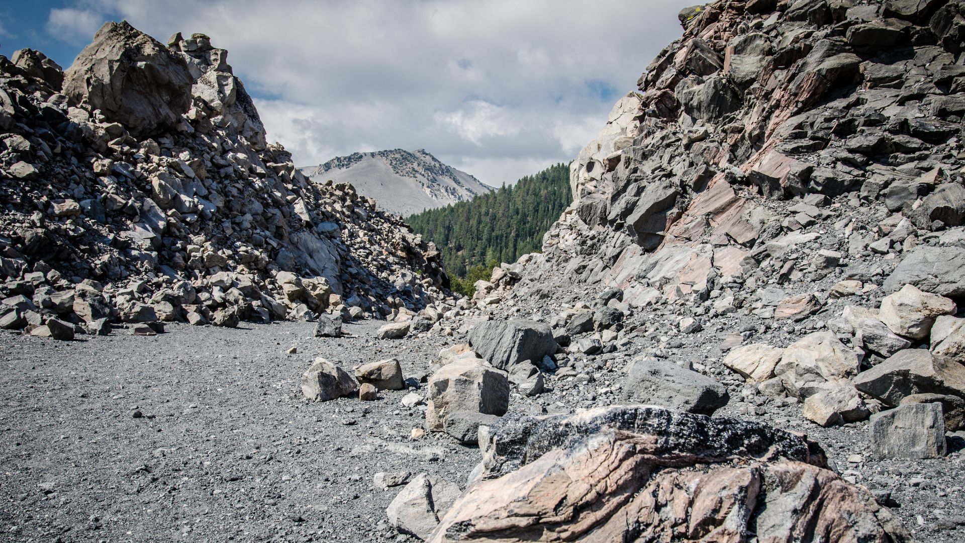 A wide shot of Obsidian Dome in California, featuring a rocky, barren landscape with large piles of dark volcanic rock and obsidian fragments, framed by steep, rocky slopes and a distant tree-covered mountain under a cloudy sky.