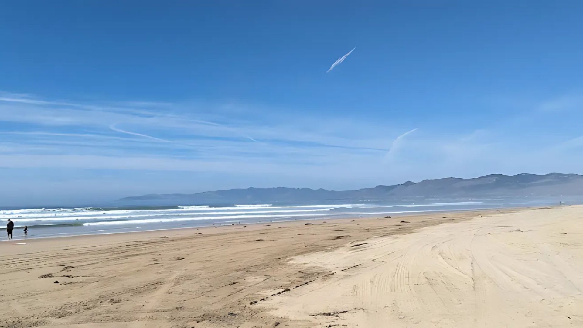 A wide-angle shot of a sunny beach with gentle waves, a vast expanse of sand, and distant mountains under a blue sky with scattered clouds and faint contrails. A few people are visible near the waterline.