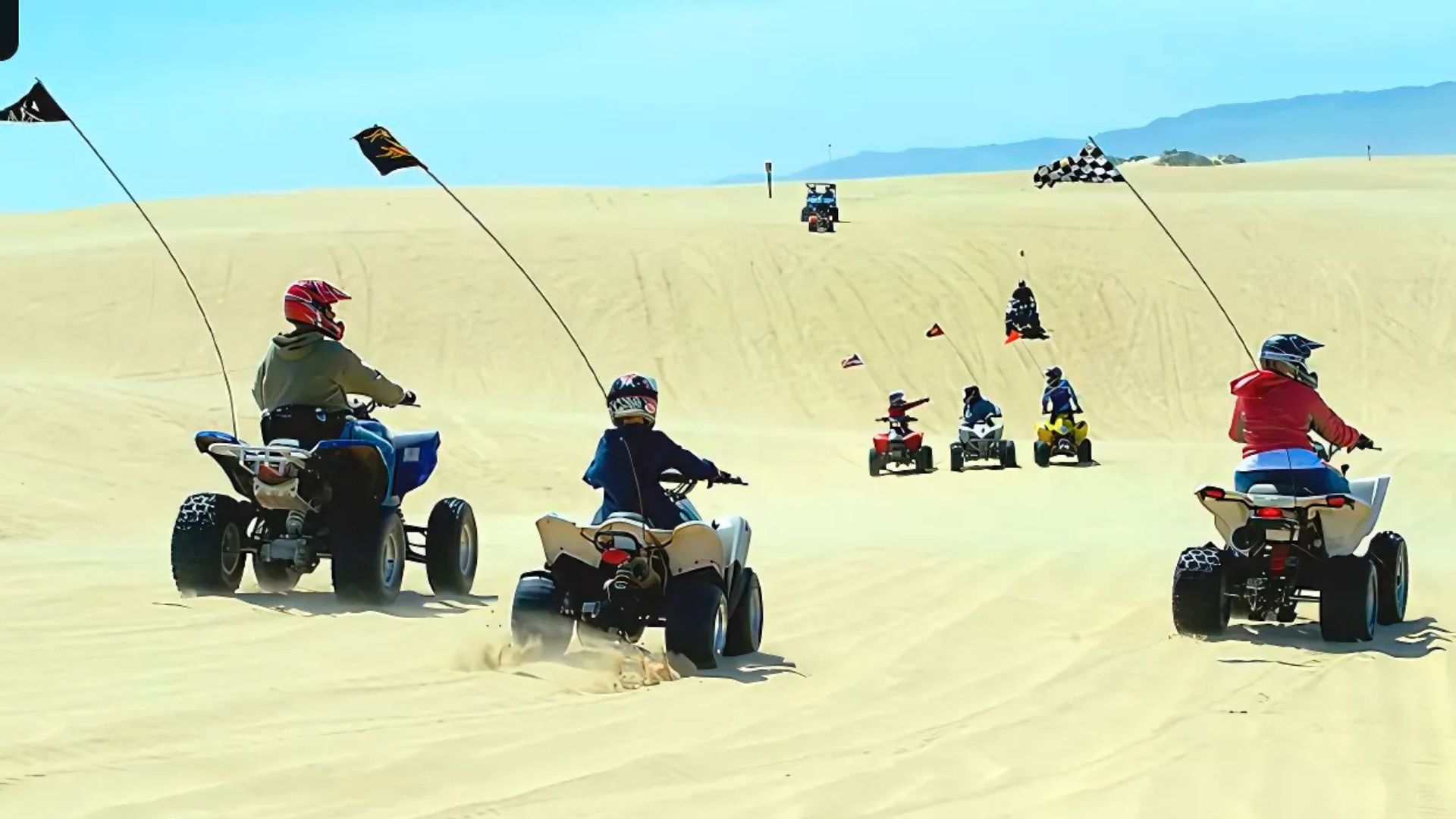 A group of people are riding all-terrain vehicles (ATVs) on large sand dunes under a clear blue sky, with some ATVs displaying flags for visibility.